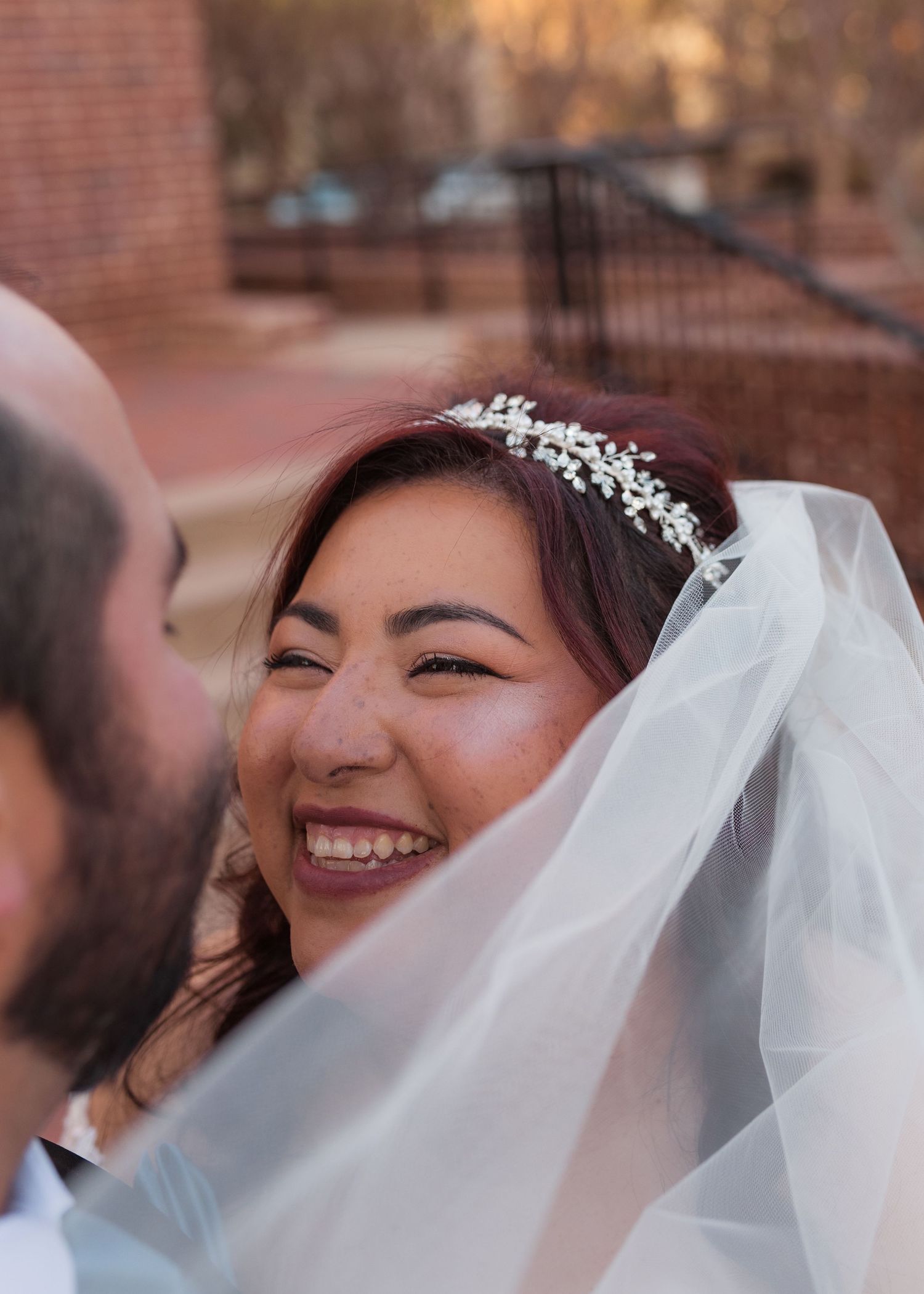 A joyful bride in a white veil and floral tiara laughs radiantly during her wedding ceremony outdoors.