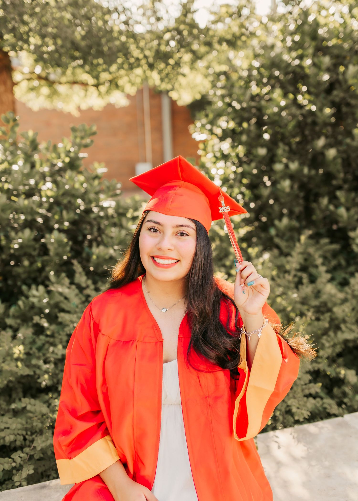 Graduate in bright red cap and gown posing outdoors against green foliage.