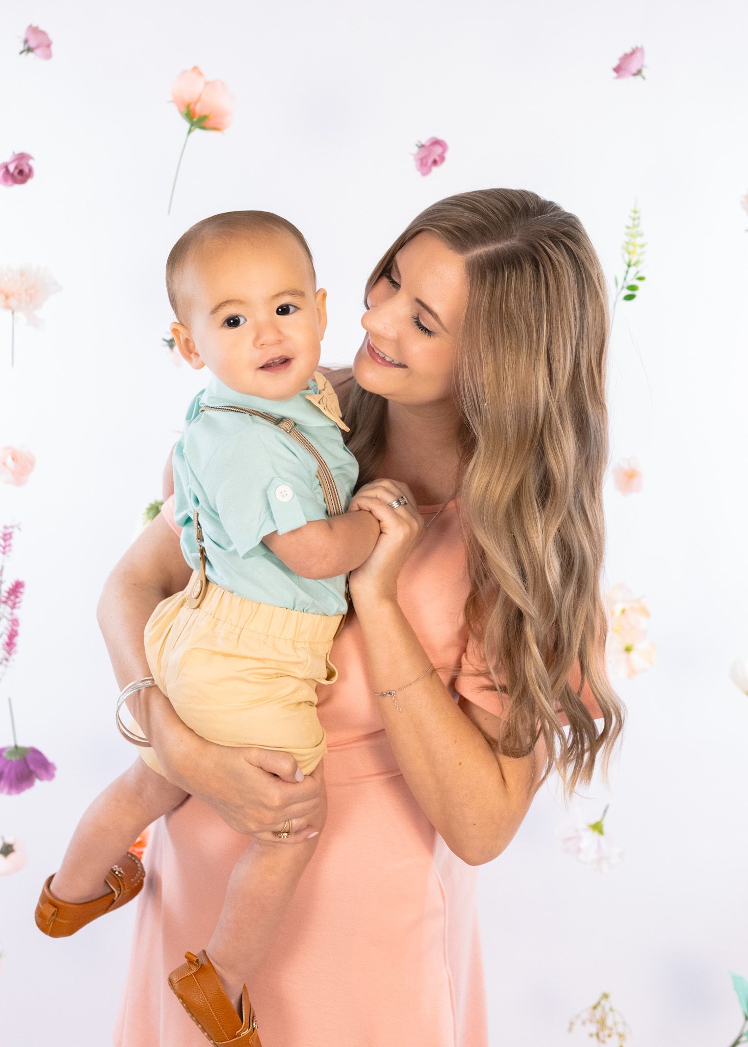 Sweet studio portrait with floral backdrop showing affectionate moment with baby in yellow shorts.