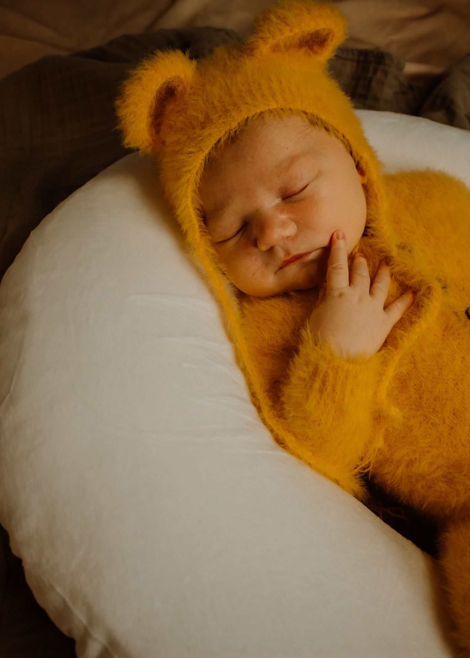 A sleeping infant wearing a cozy yellow bear onesie with furry ears lies on a white pillow.