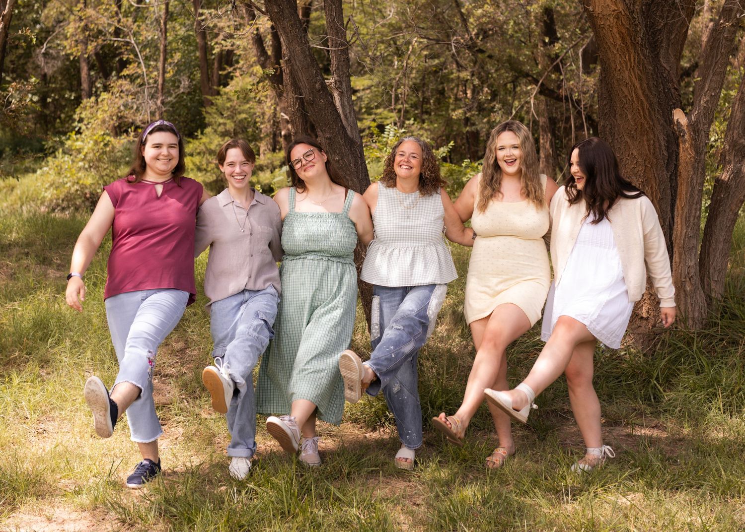 Six women laughing and posing together outdoors in a sunny forest setting, wearing colorful summer outfits.