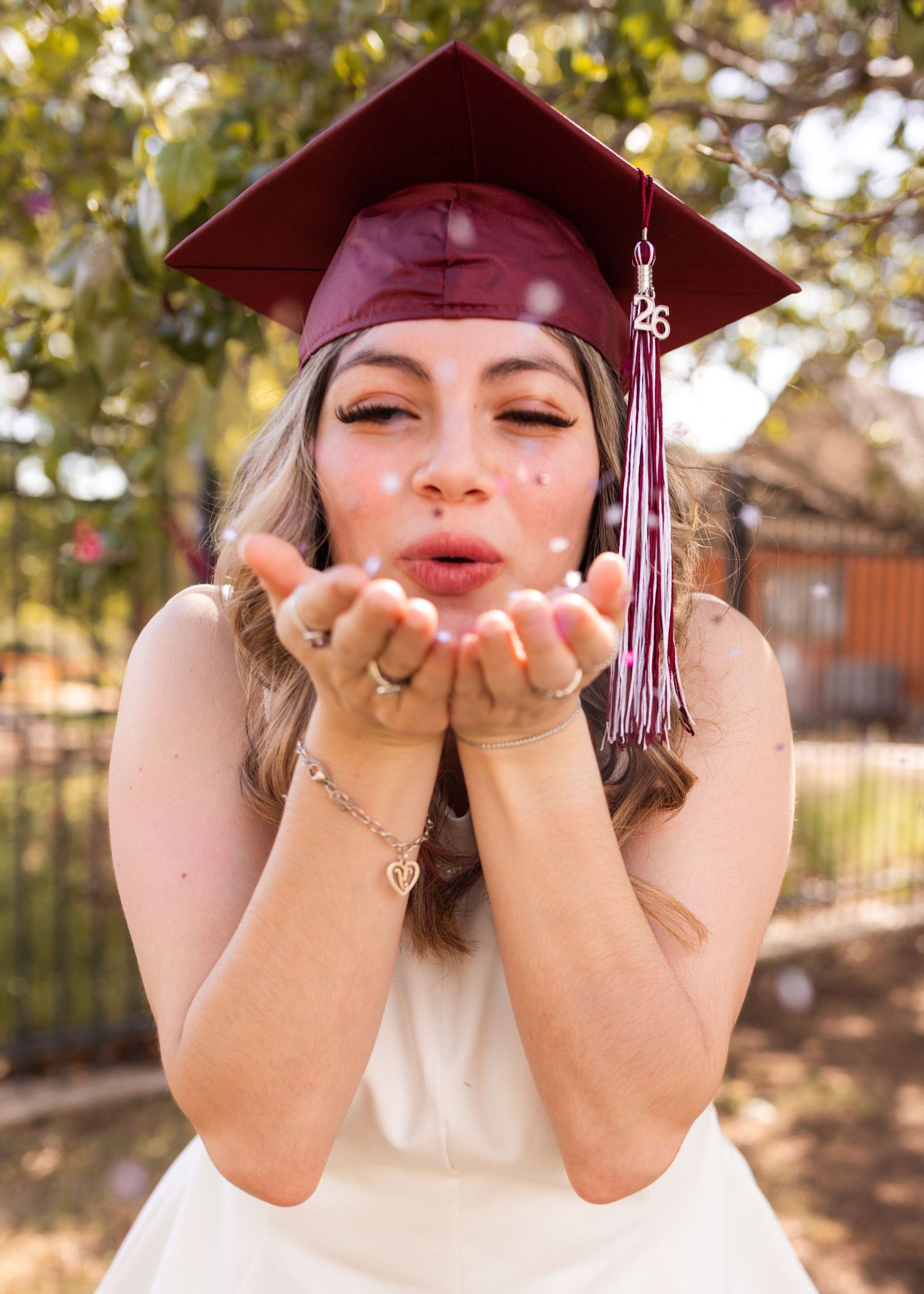 Graduate in maroon cap blowing glitter from her hands outdoors, celebrating her 2023 graduation.