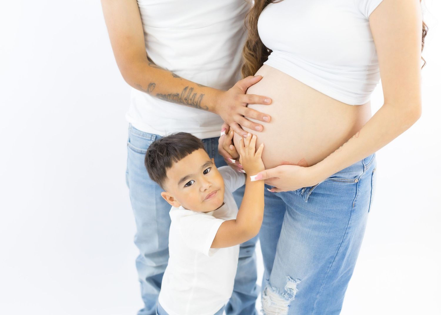 Young boy hugging pregnant mother's belly while standing with parents against white background.