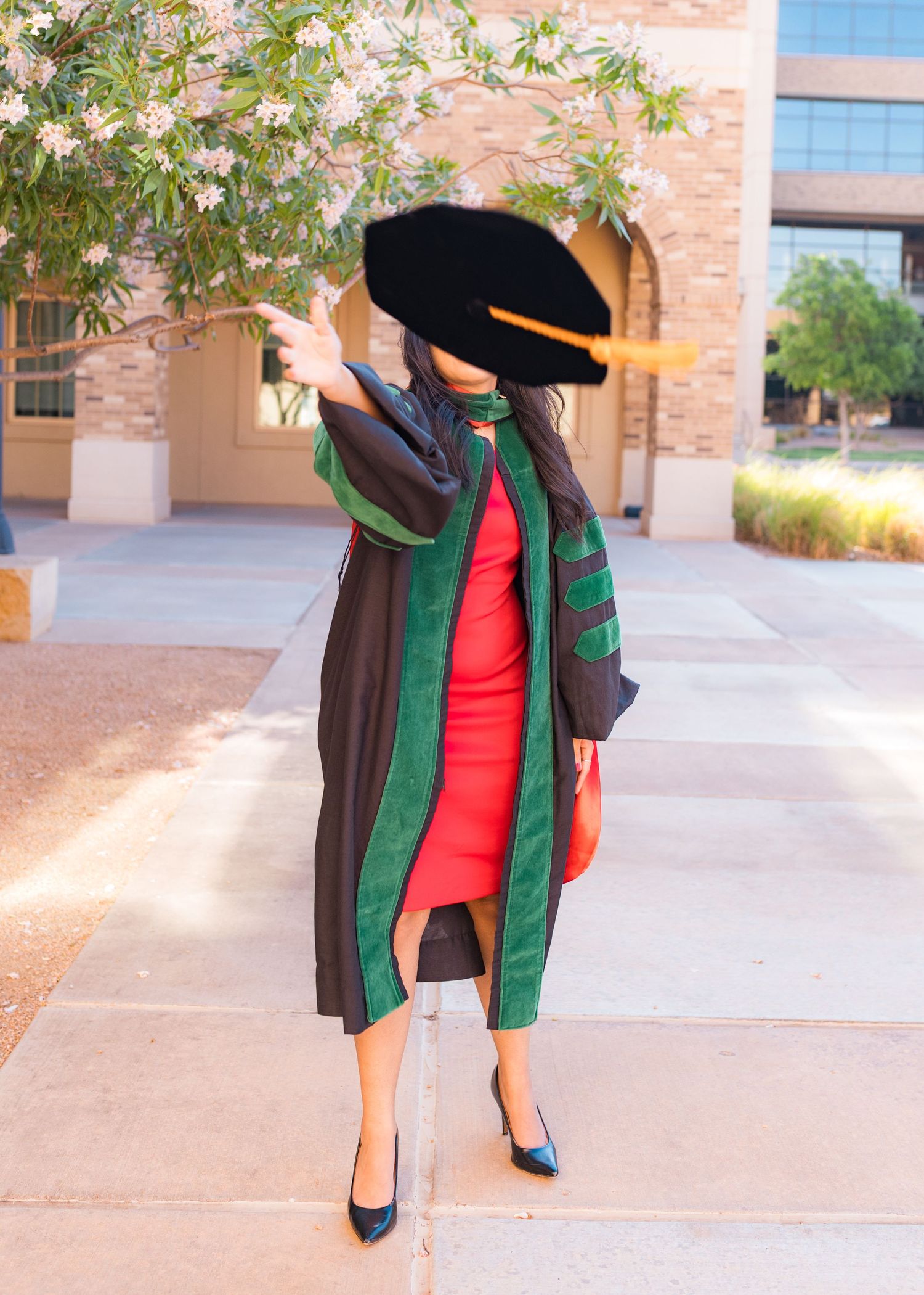 Graduate in black doctoral gown, green hood, and tam cap poses outdoors near flowering tree in red dress.