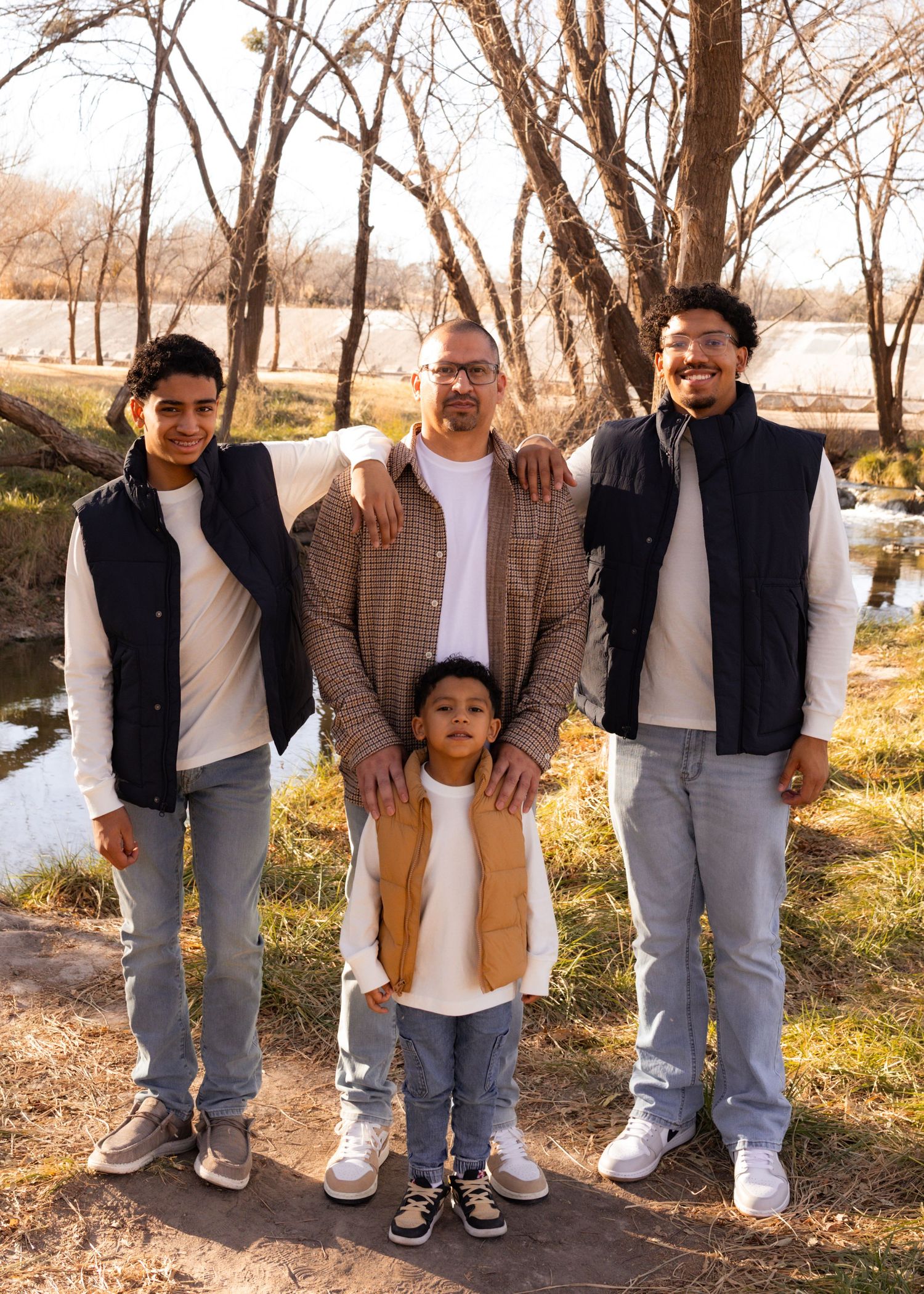 A family of four poses outdoors near a lake with bare trees, wearing casual layered outfits in a winter setting.