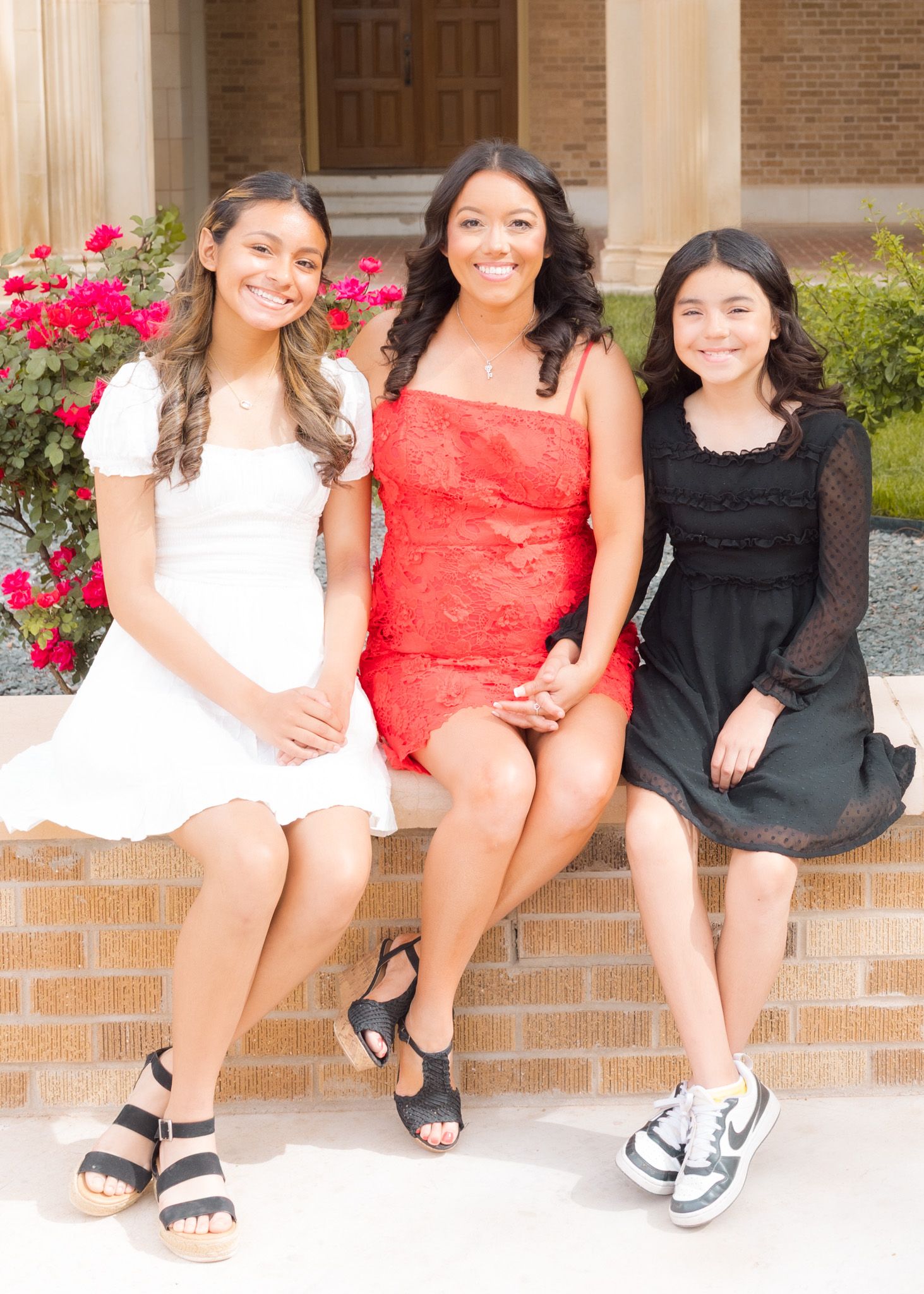 Three people in dresses sit on brick steps with red flowers blooming in the background.