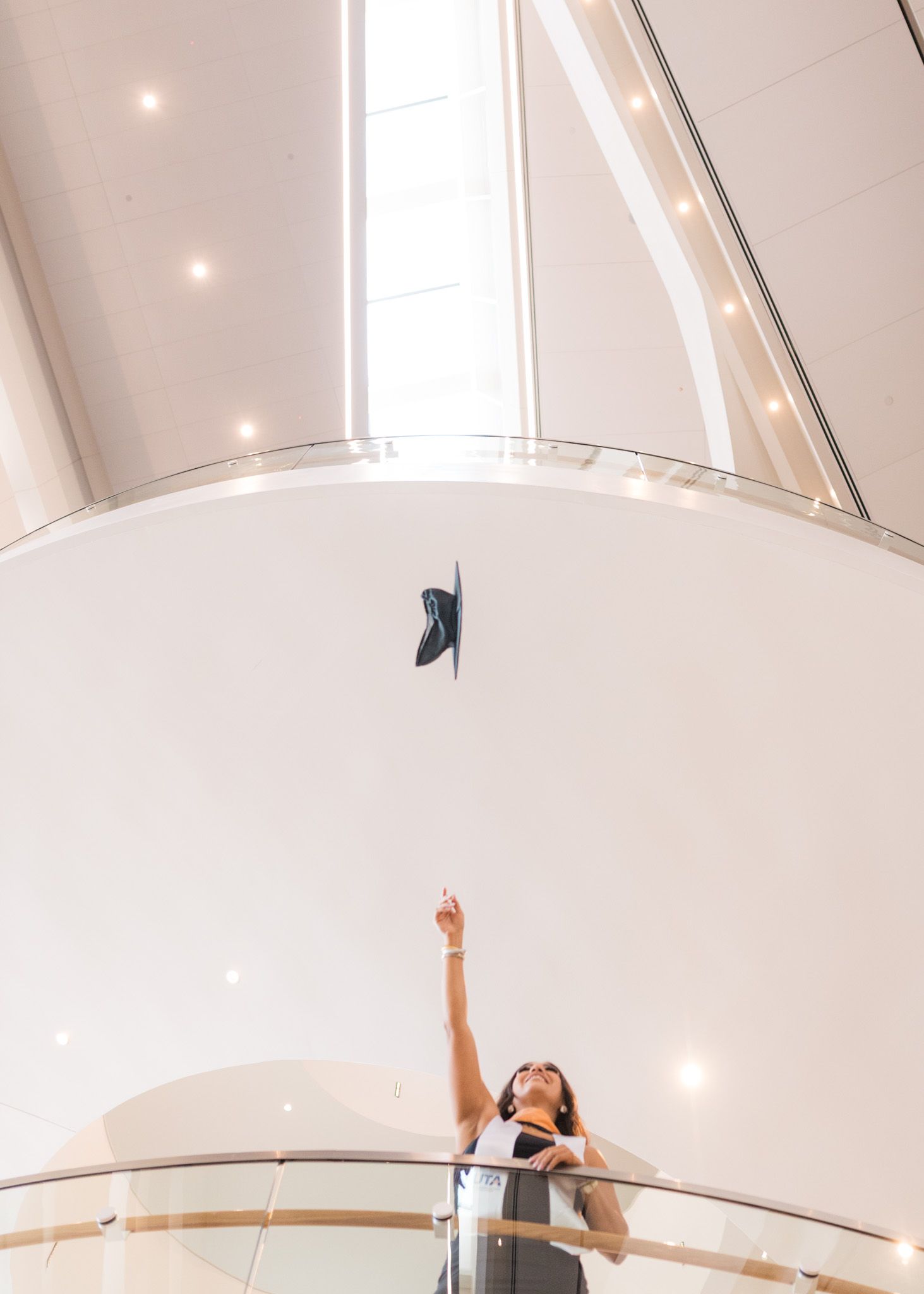 Graduate tosses cap in celebration on modern spiral staircase with white walls.
