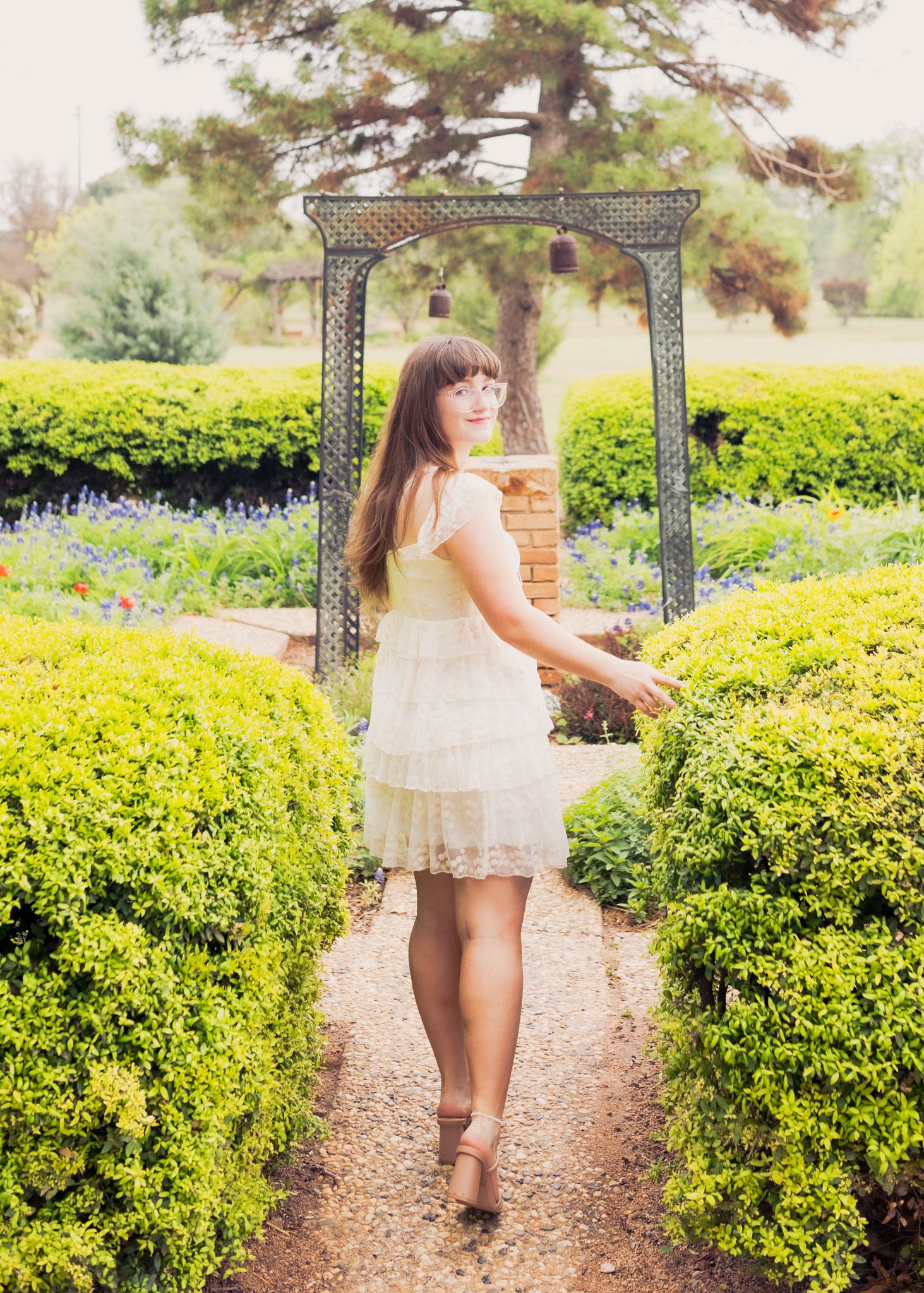 White sundress and garden path leading to decorative metal arbor surrounded by manicured hedges.