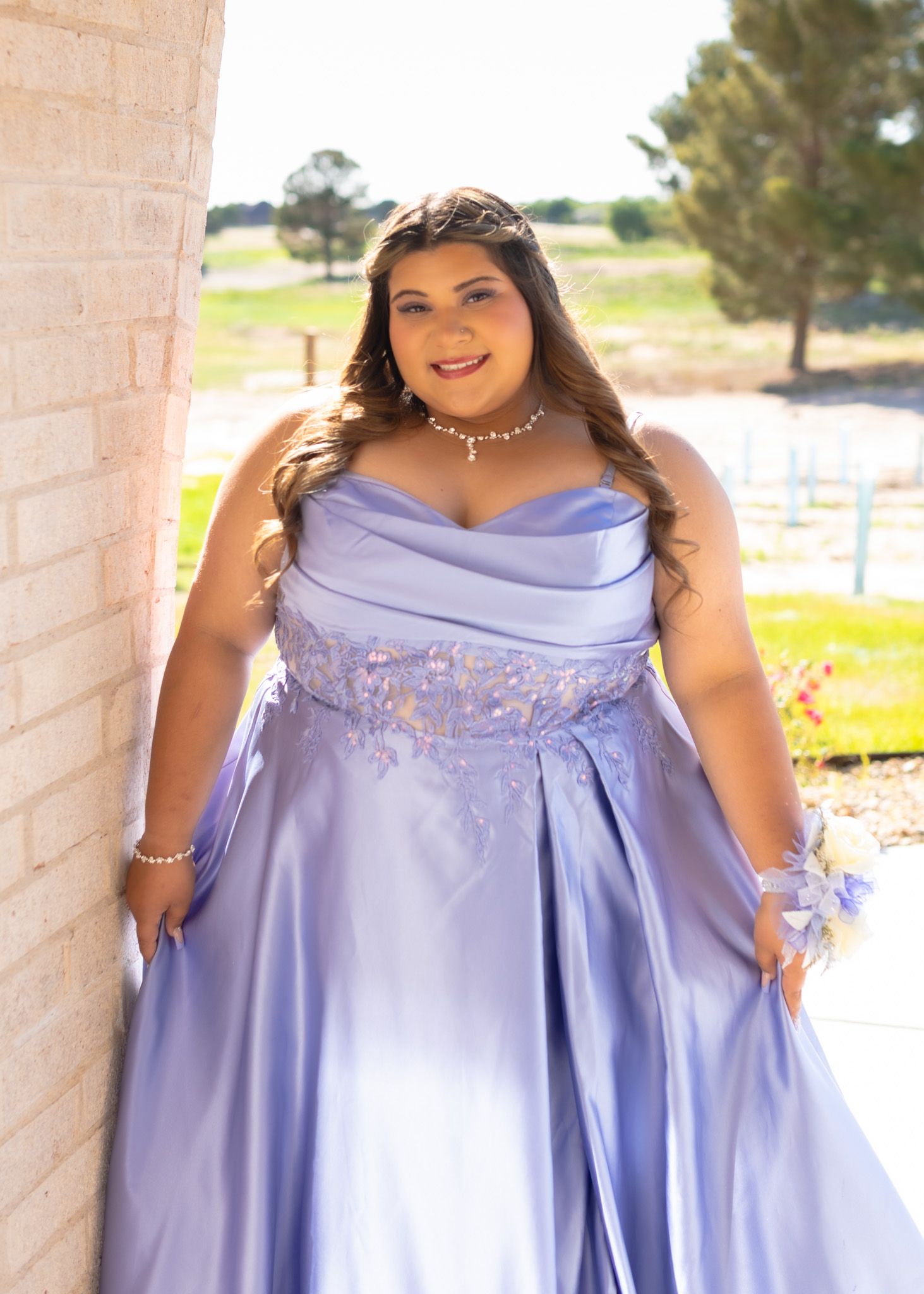 A plus-size model in a lavender satin gown with beaded detailing poses against a light-colored wall outdoors.