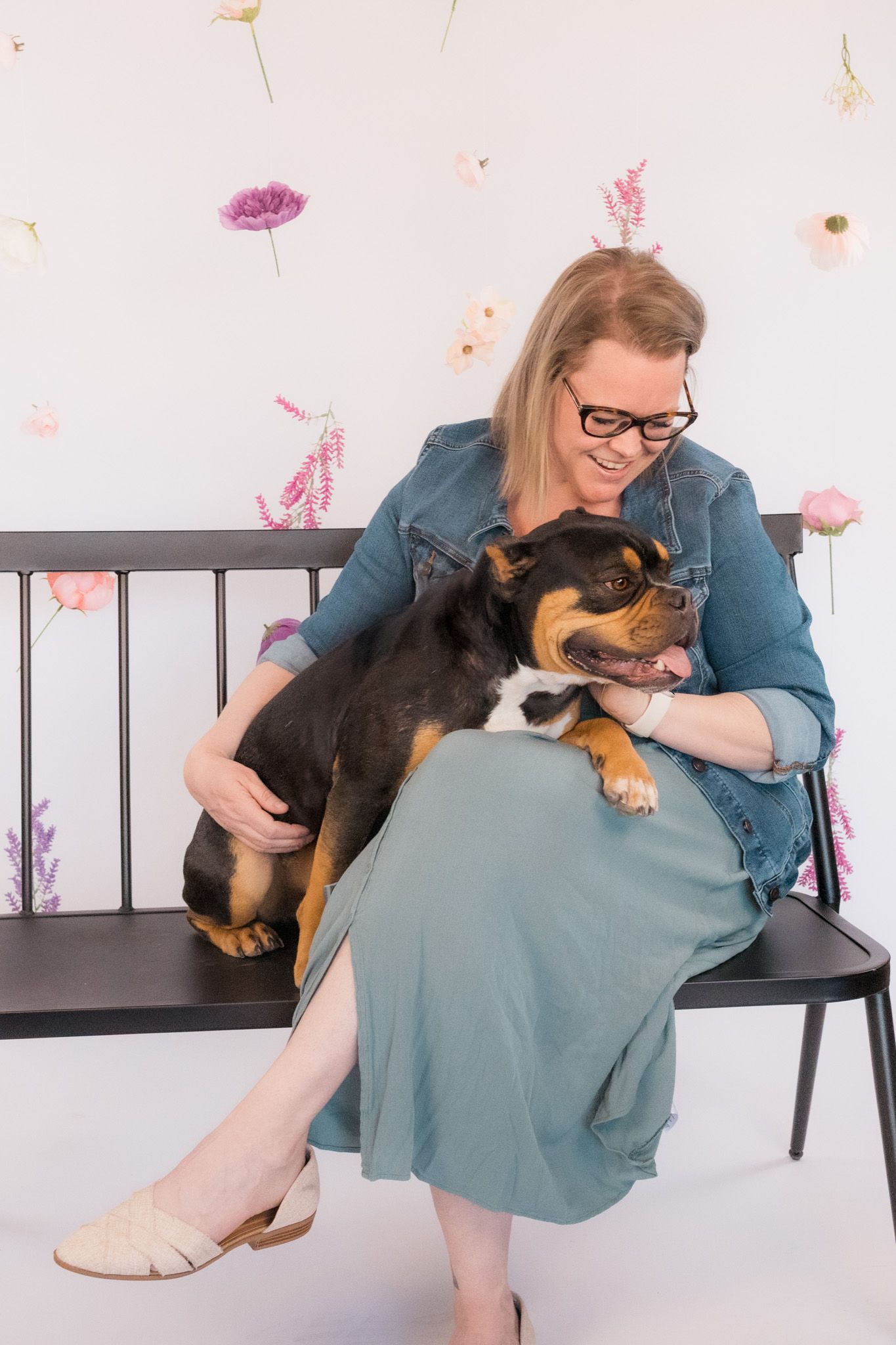 Woman in denim jacket cuddles with mixed breed dog on black bench against floral wall.
