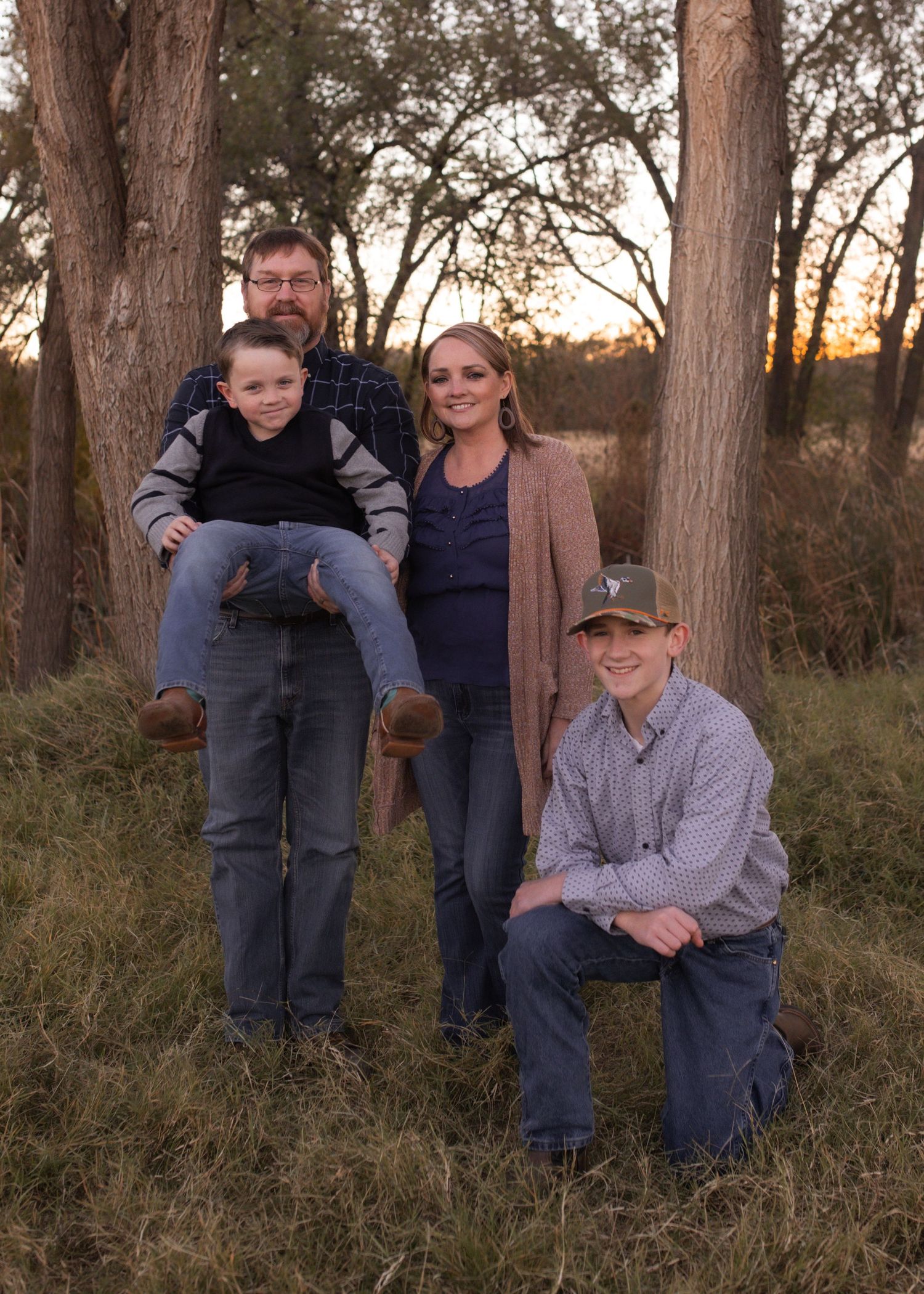 Family of four posing outdoors at sunset among tall trees, with father holding young boy and teenager kneeling in front.