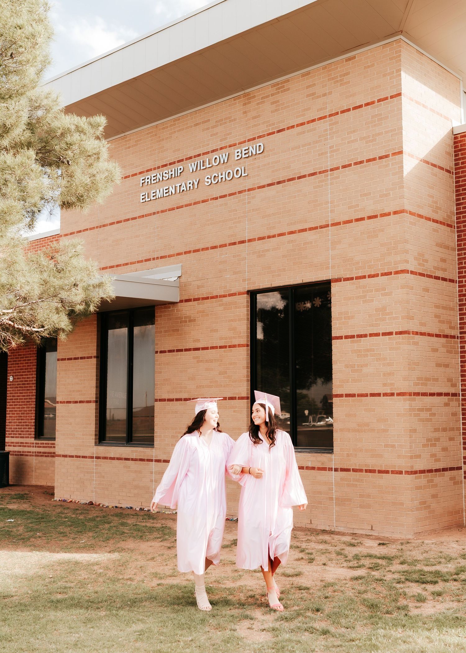 Two graduates in light pink gowns walk together outside a brick elementary school building on a sunny day.