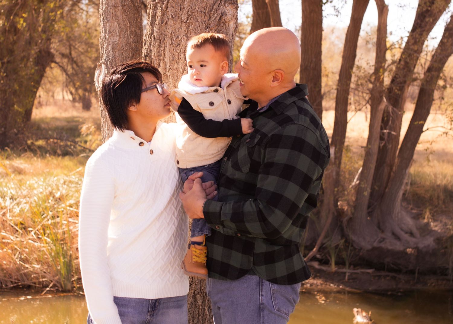 Family portrait of two parents holding their baby outdoors among autumn trees near a stream.
