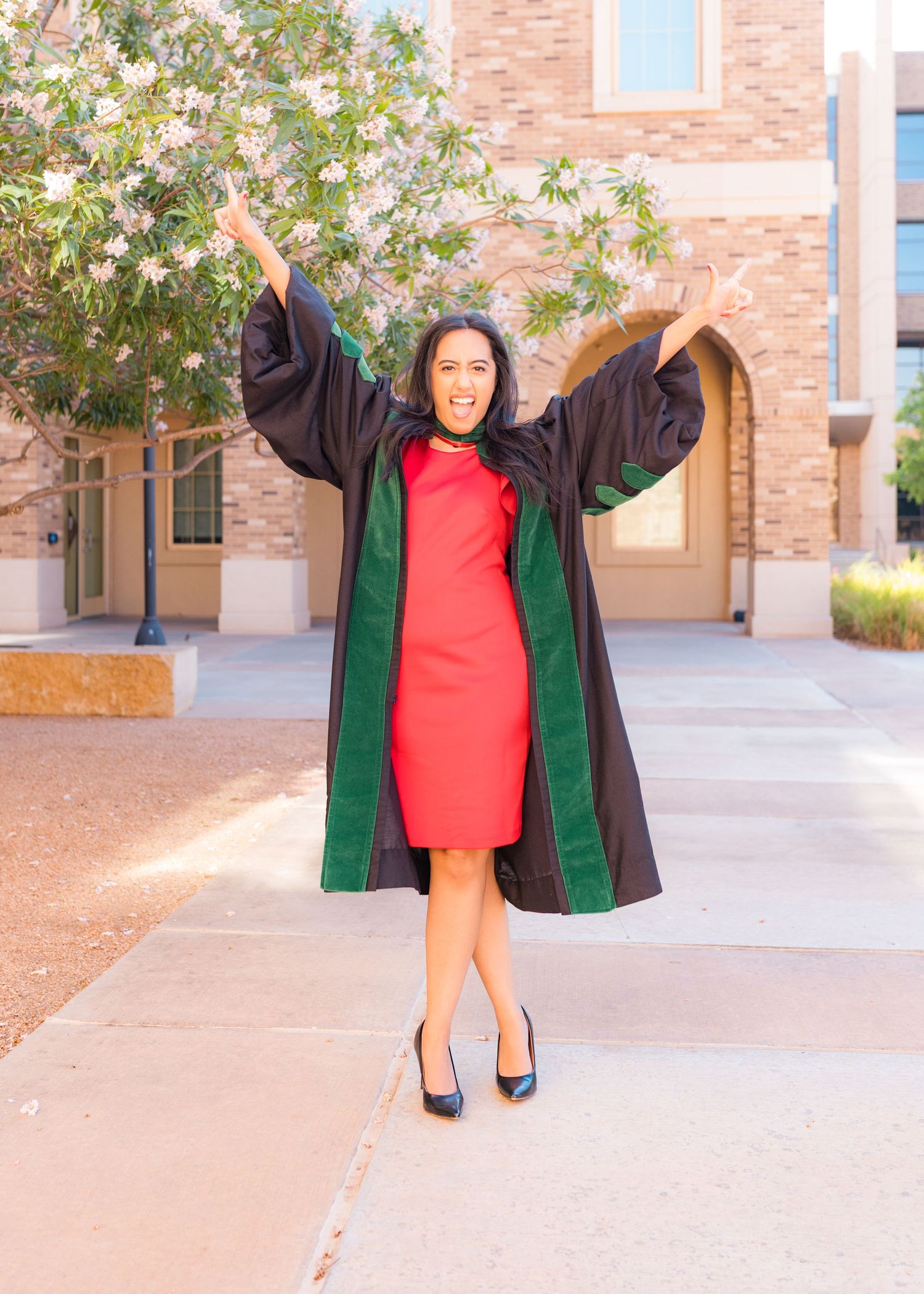 Woman in graduation gown with green velvet trim celebrates outdoors, arms raised joyfully, wearing red dress and heels.