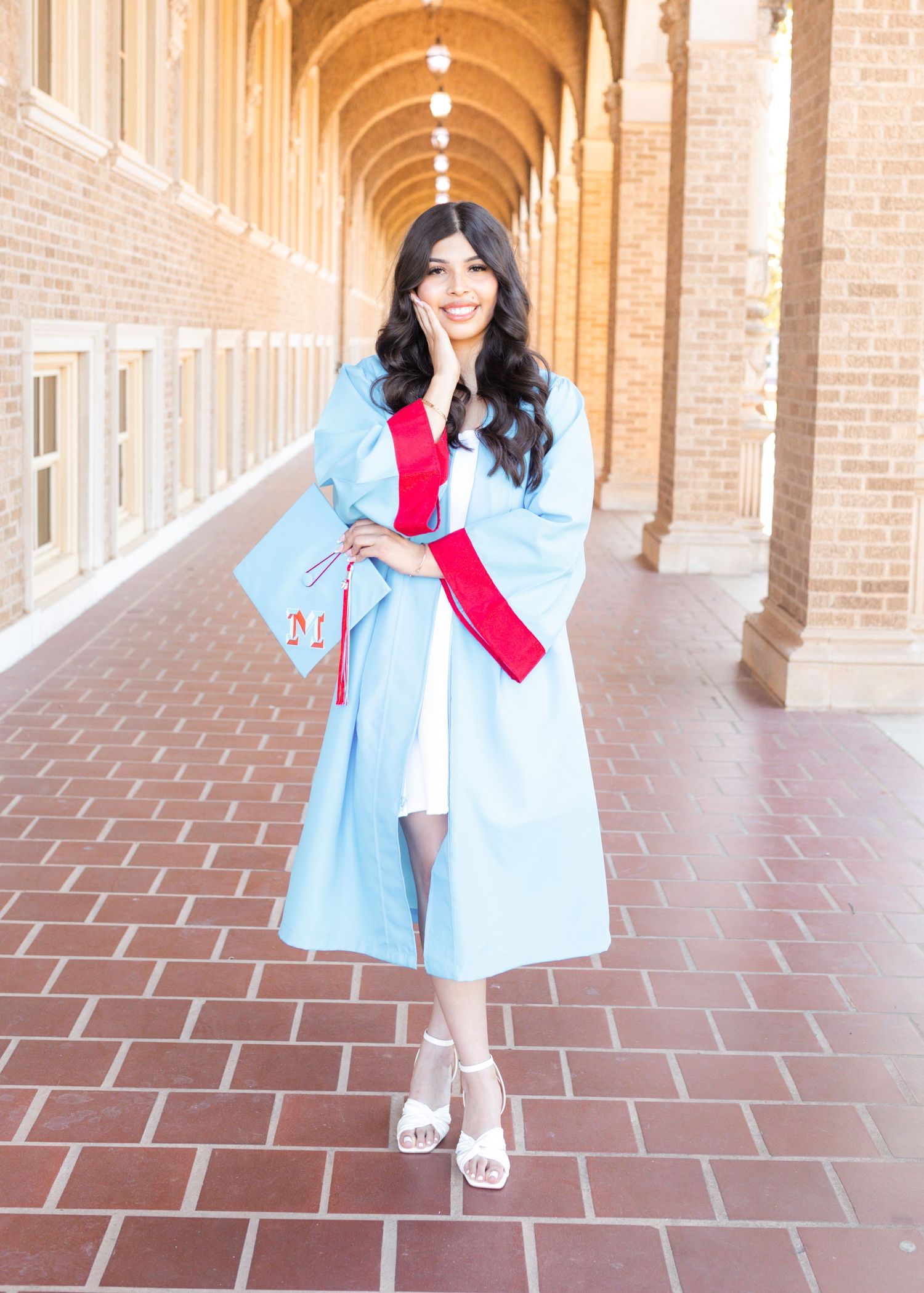 A young woman in a light blue graduation gown with red sash poses in a brick archway corridor holding her cap.