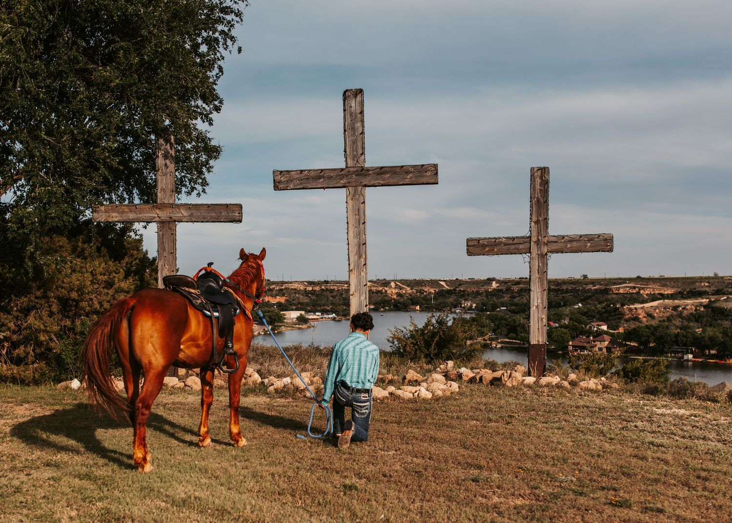 Person with horse stands reverently before three wooden crosses on hilltop at sunset.