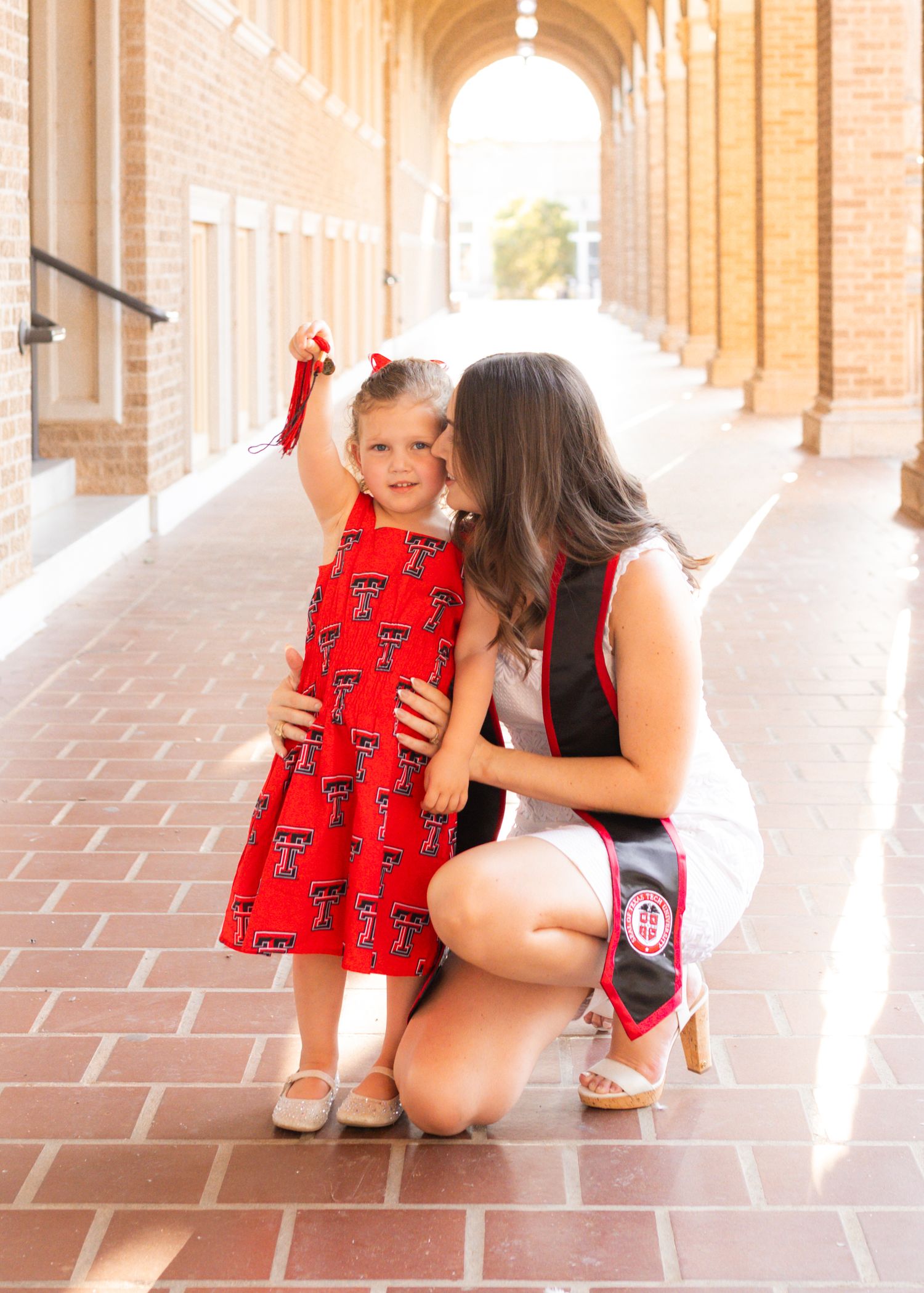 A woman in graduation attire kisses a toddler girl in a red Texas Tech dress on a brick campus walkway.