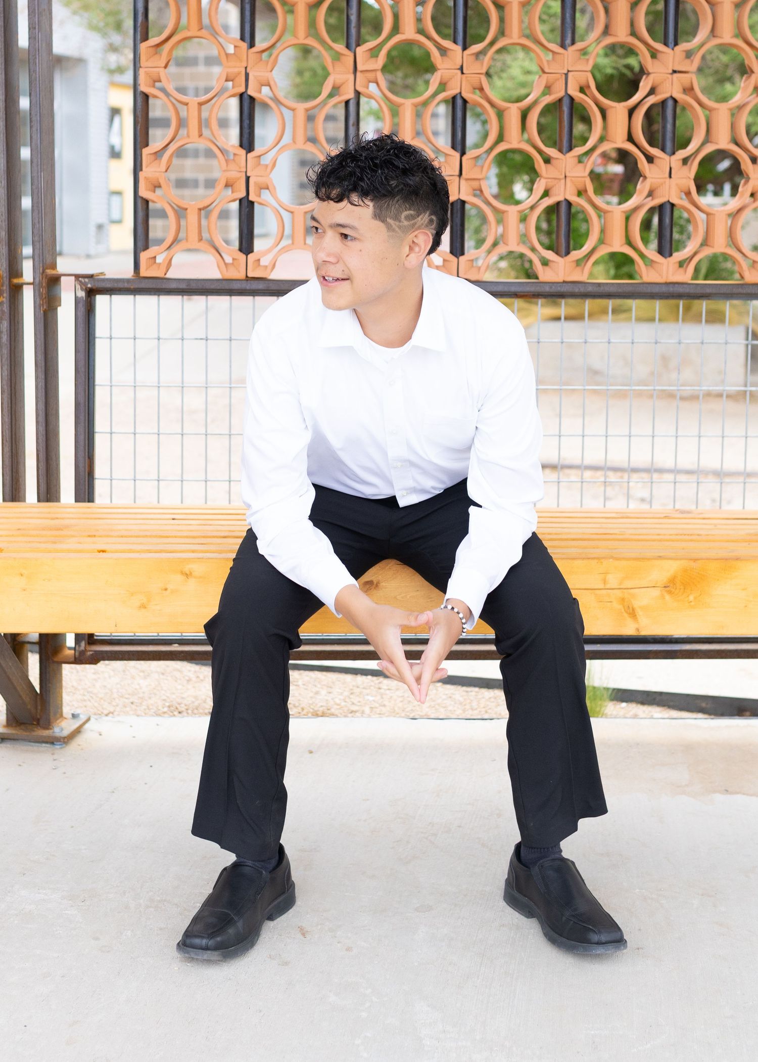 Young man in white shirt and black pants sitting on a wooden bench outdoors, smiling confidently.