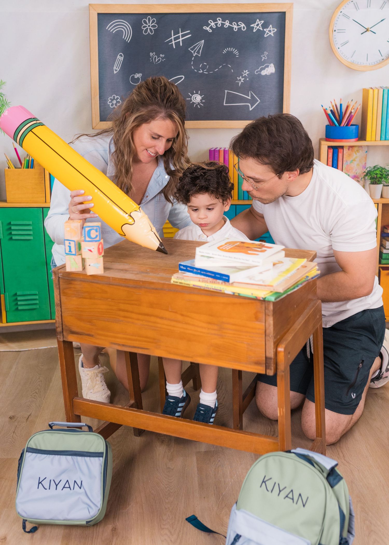 Two parents help young child named Kiyan study at wooden desk with giant pencil prop and chalkboard backdrop.