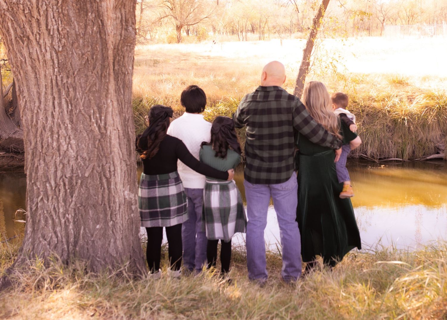 Family of six standing together by a large tree near a peaceful pond in a scenic outdoor autumn setting.