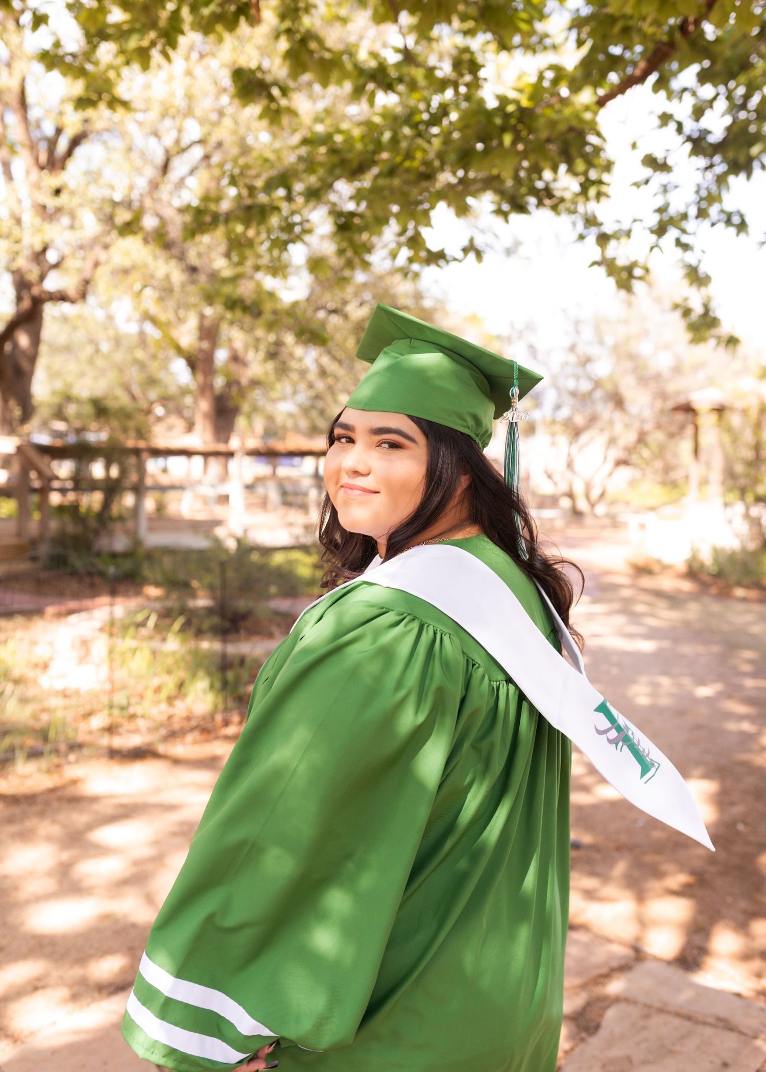 Young woman in green graduation cap and gown smiles over her shoulder outdoors among trees.