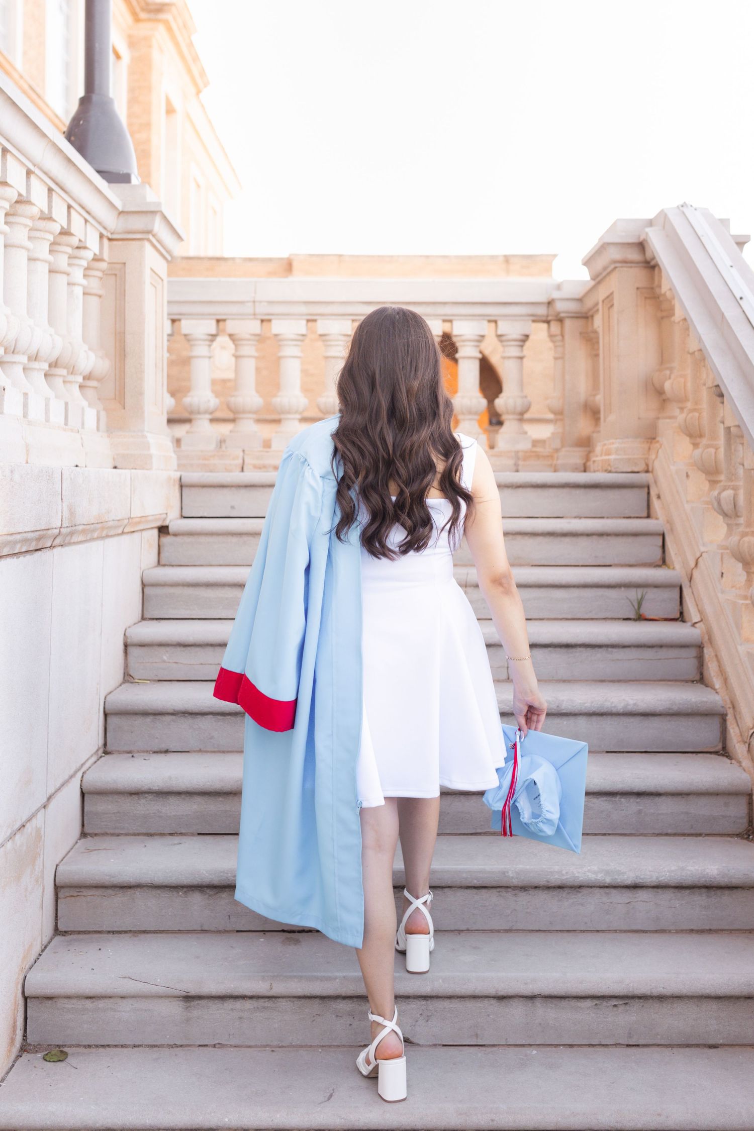 Young woman in white dress and blue graduation gown ascending stone steps, holding cap, viewed from behind.