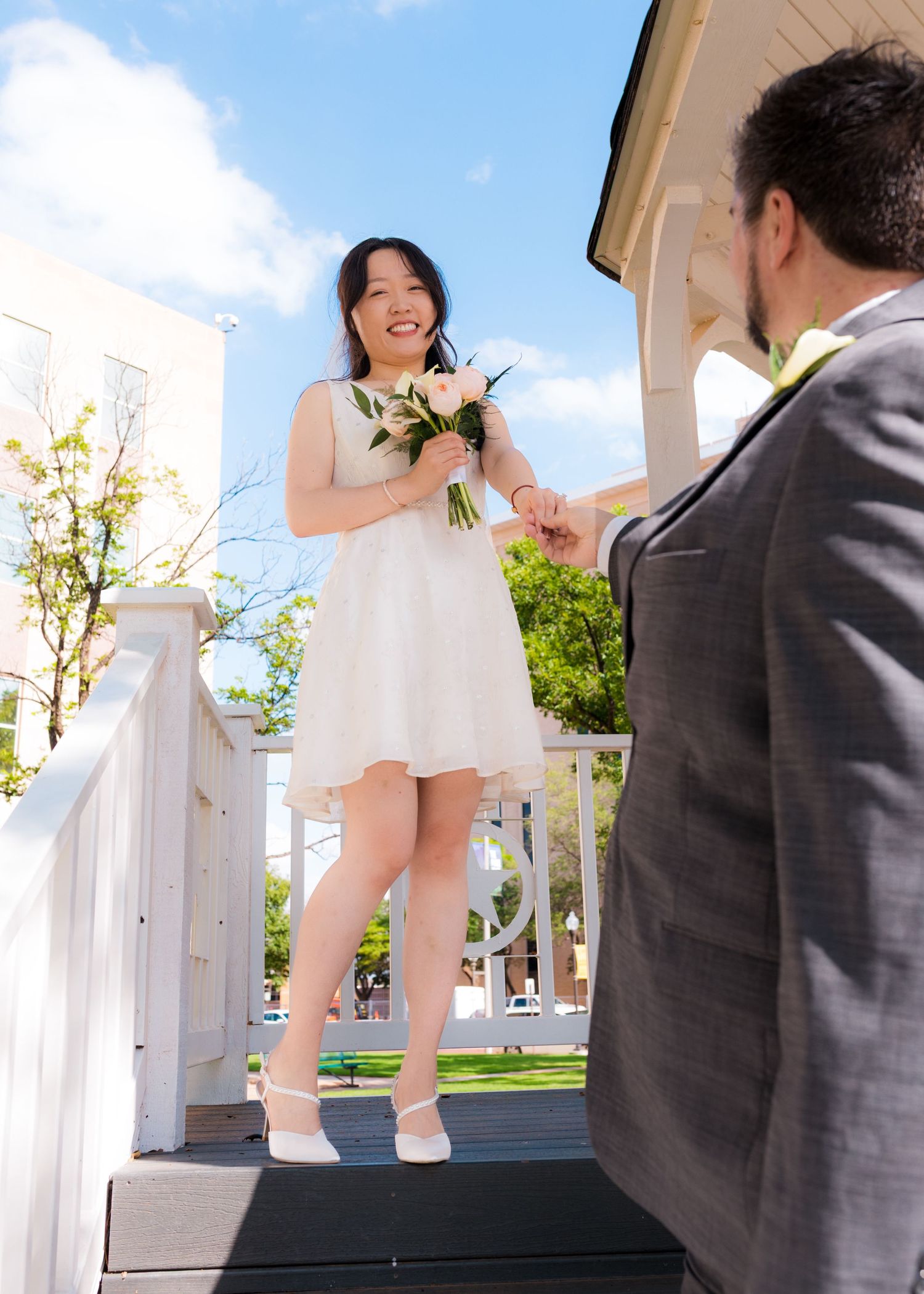 Smiling Asian bride in white dress holding bouquet exchanges hands with groom in gray suit on outdoor wedding venue steps.