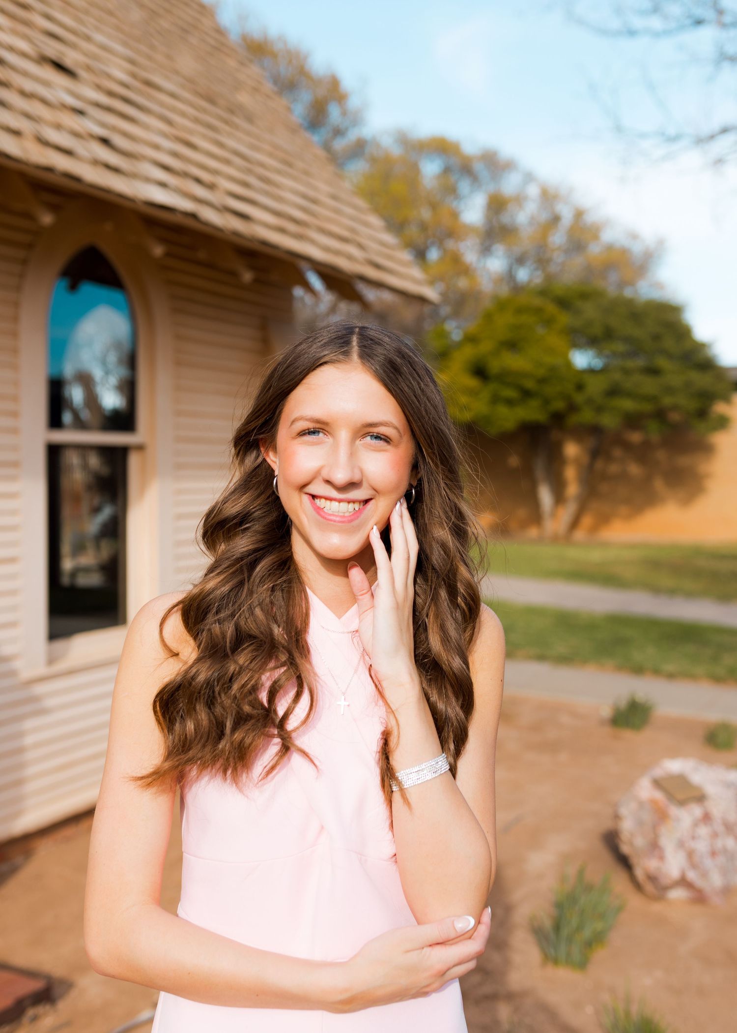Smiling young woman in pink sleeveless top standing outdoors near a rustic building on a sunny day.
