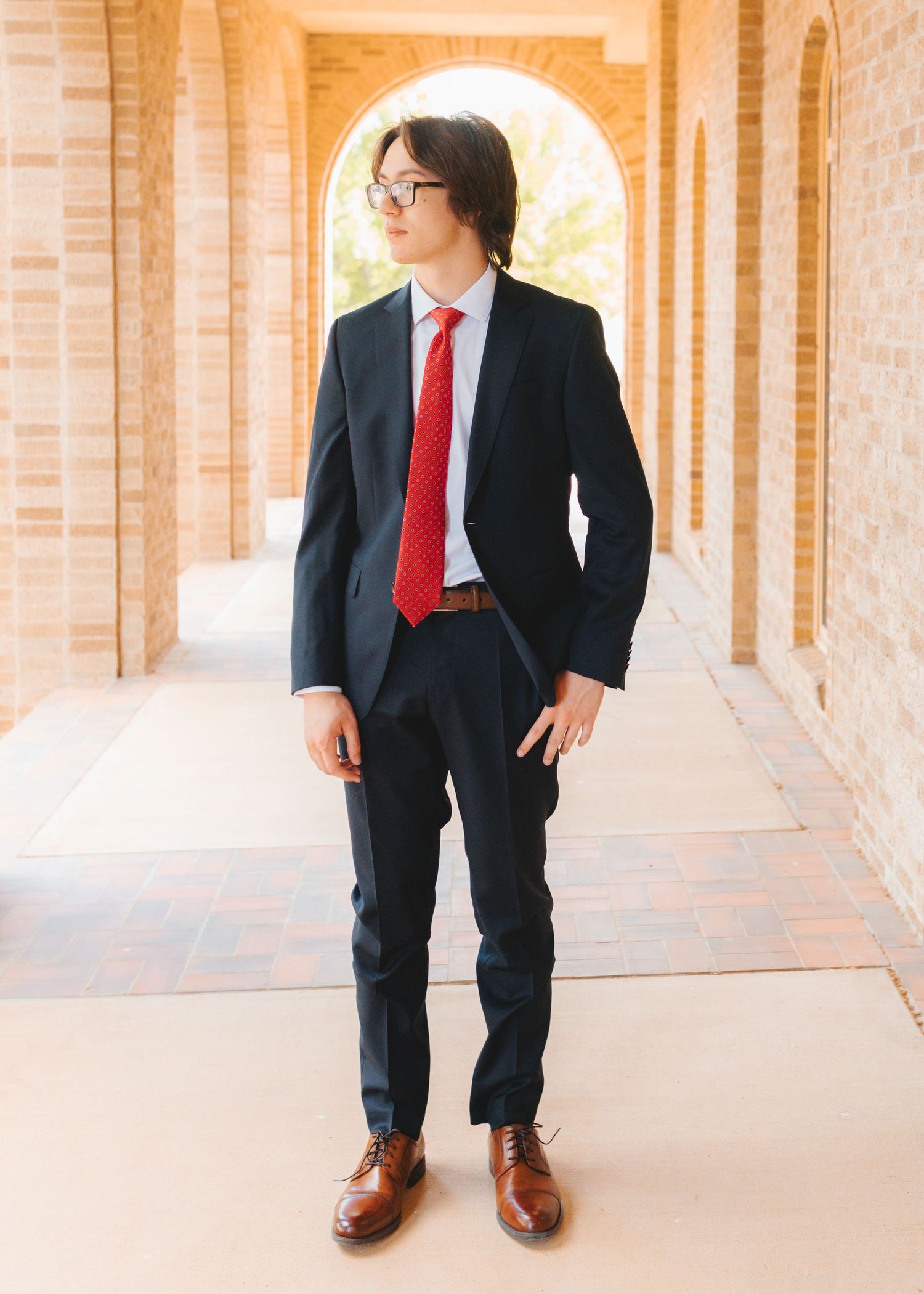 Young man in navy suit, red tie, glasses, and brown dress shoes standing in brick archway corridor.