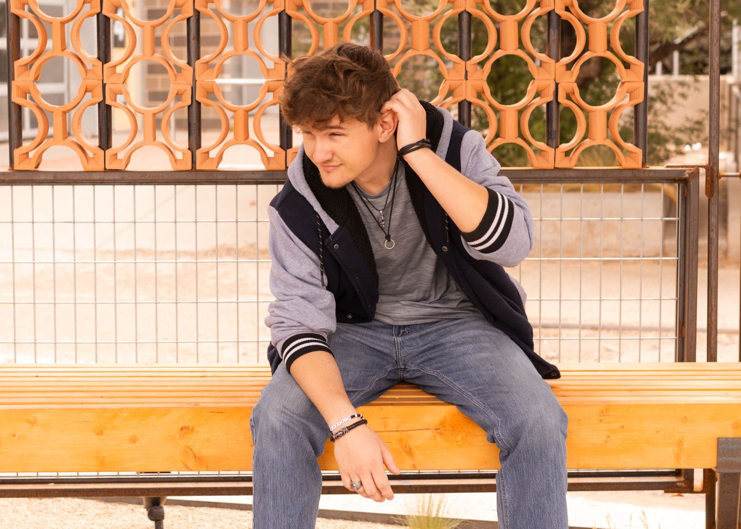 Teenage boy in gray hoodie and jeans sitting on wooden bench outdoors, looking thoughtful.