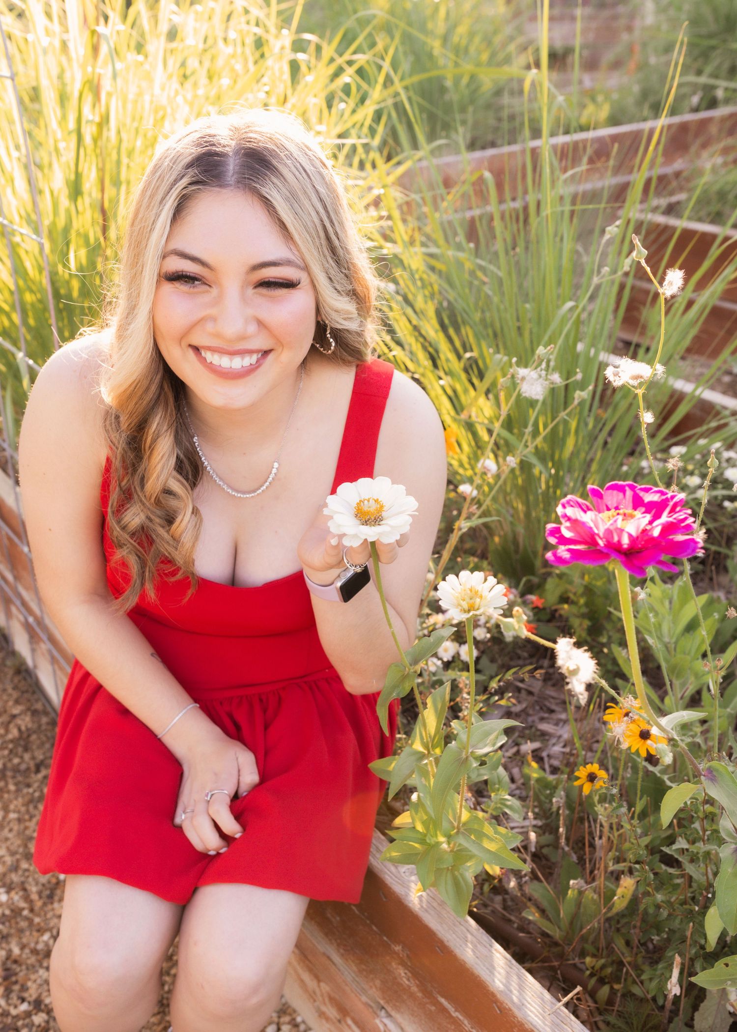 Smiling young woman in red dress holding a white daisy while sitting among colorful garden flowers outdoors.