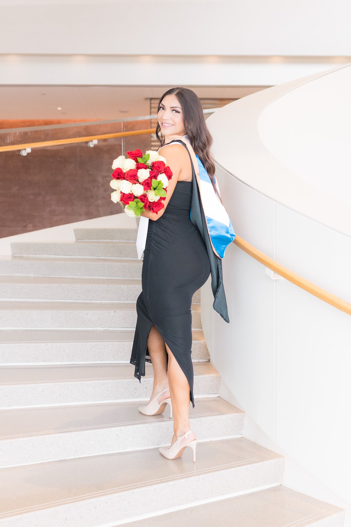 Elegant woman in black dress ascending staircase with heart-shaped bouquet of red and white roses and blue handbag.