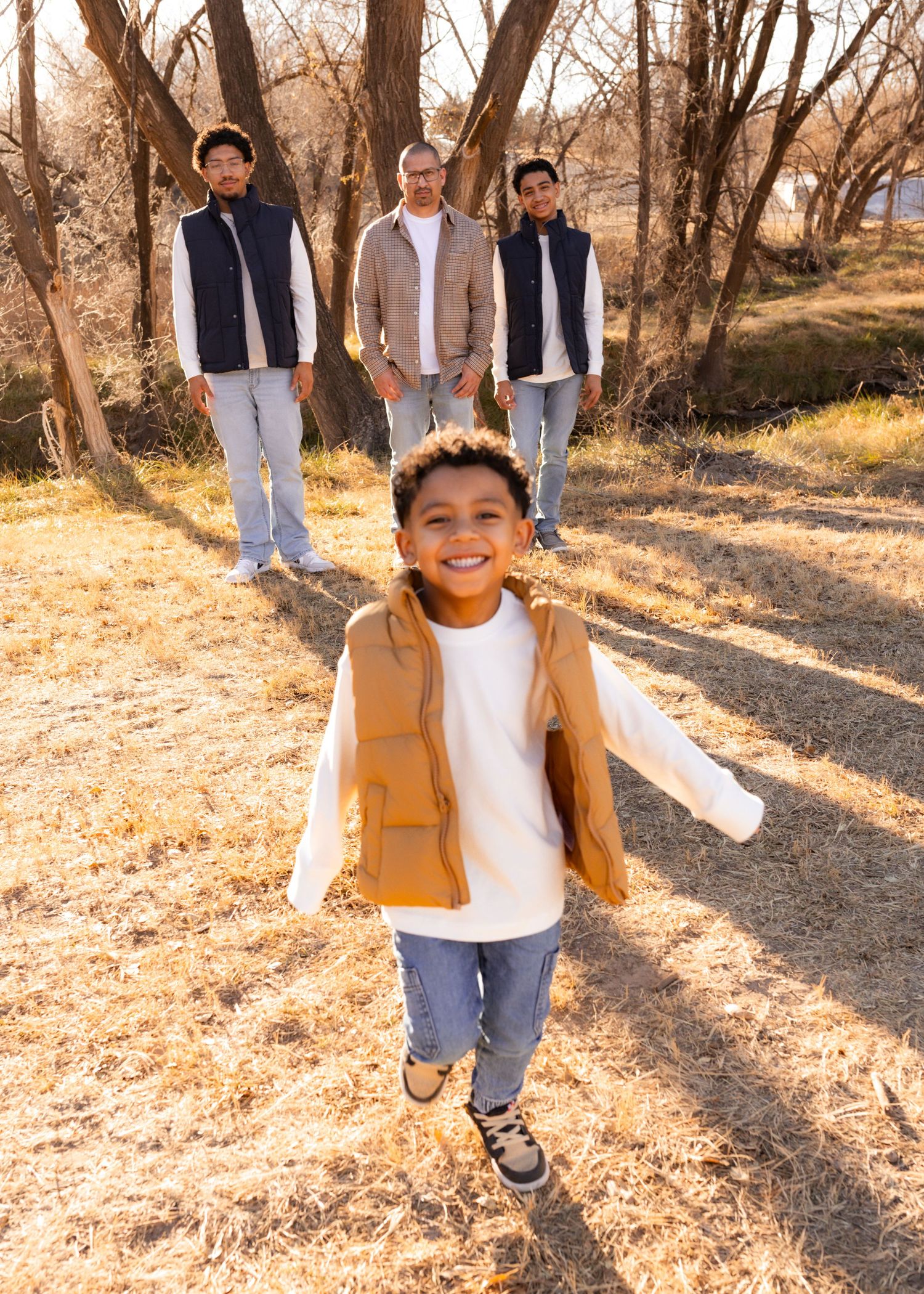 A joyful young boy in a tan vest runs toward the camera while three men stand smiling in a sunny outdoor setting.