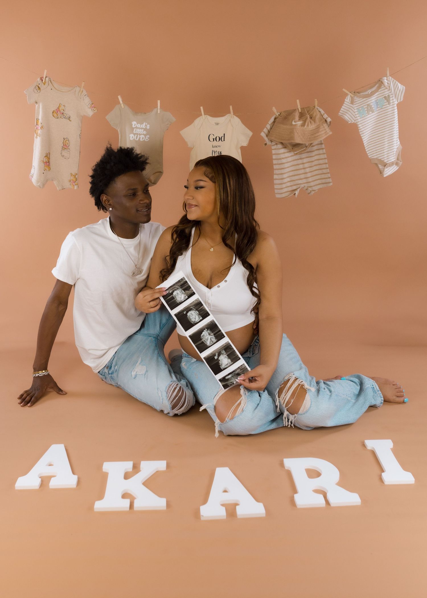 Couple sitting on floor holding ultrasound photos beside letter name AKARI with baby onesies hanging in background.