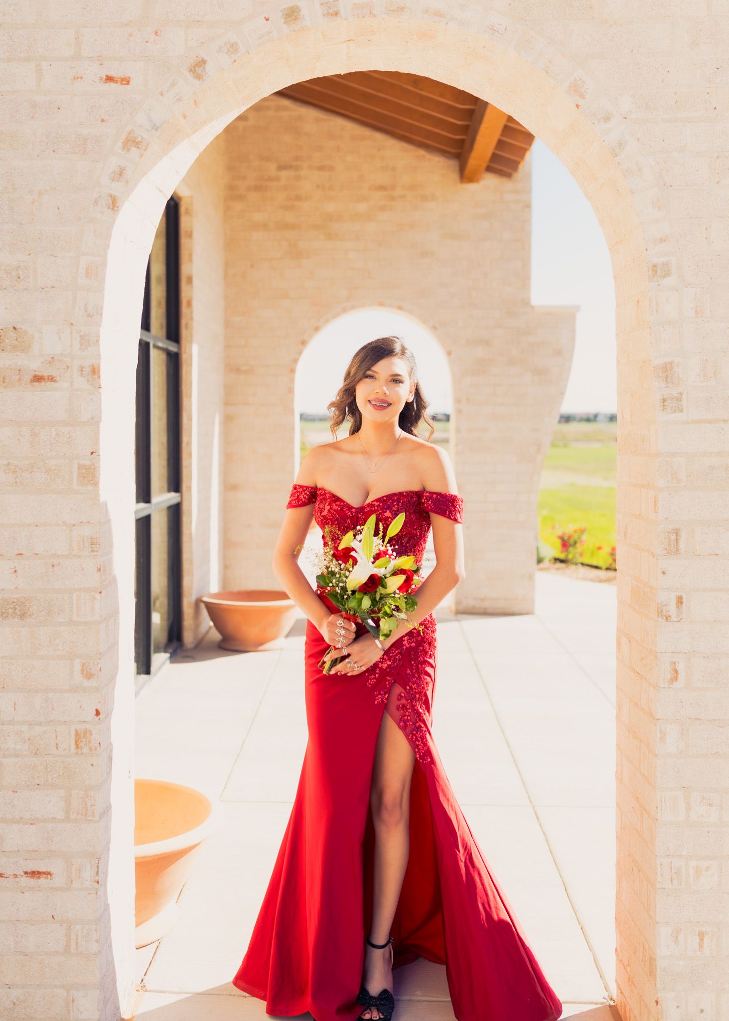 An elegant figure poses in a red off-shoulder gown with a thigh-high slit under a stone archway.