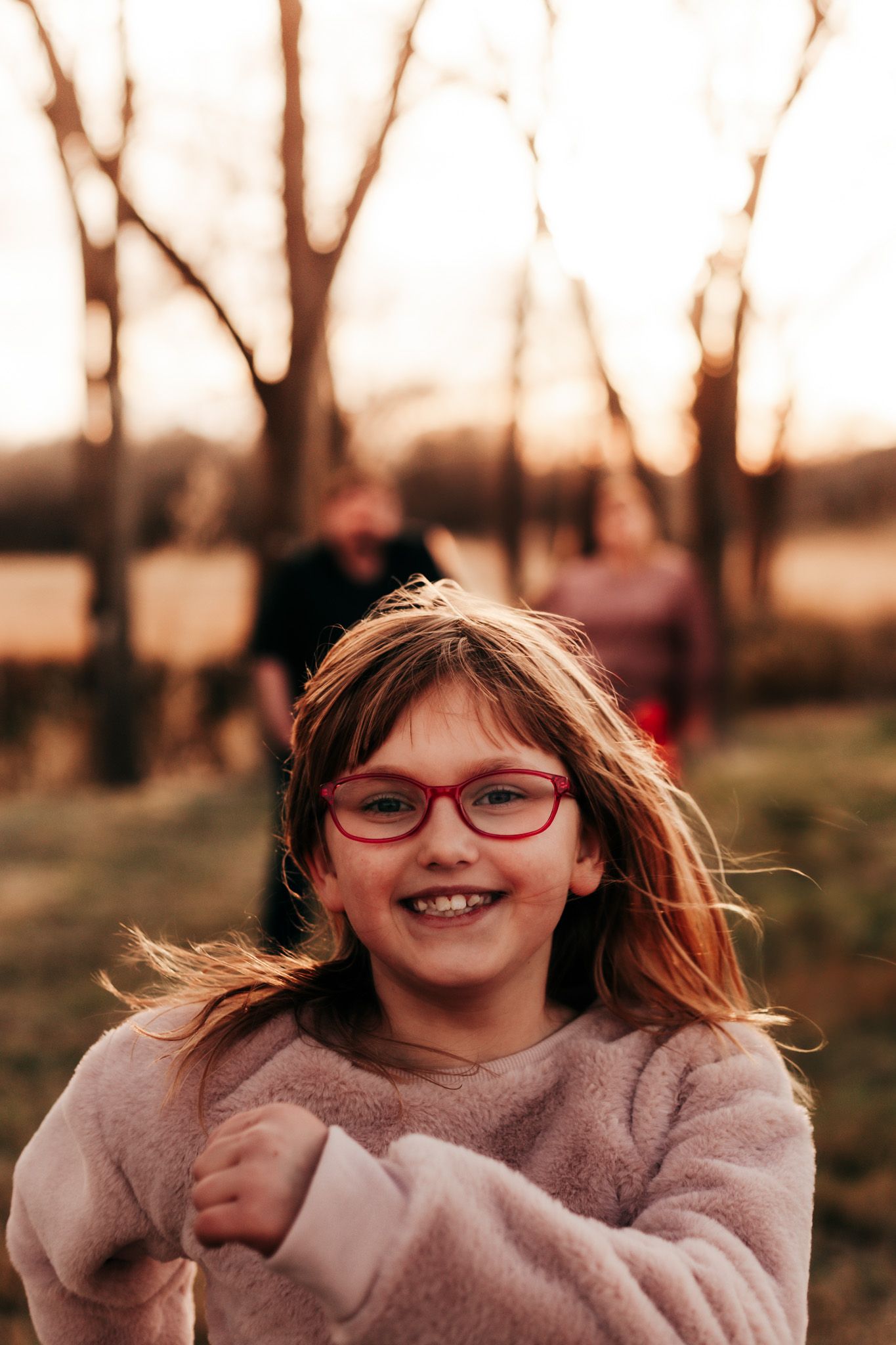 Child with red glasses smiling brightly outdoors during golden hour.