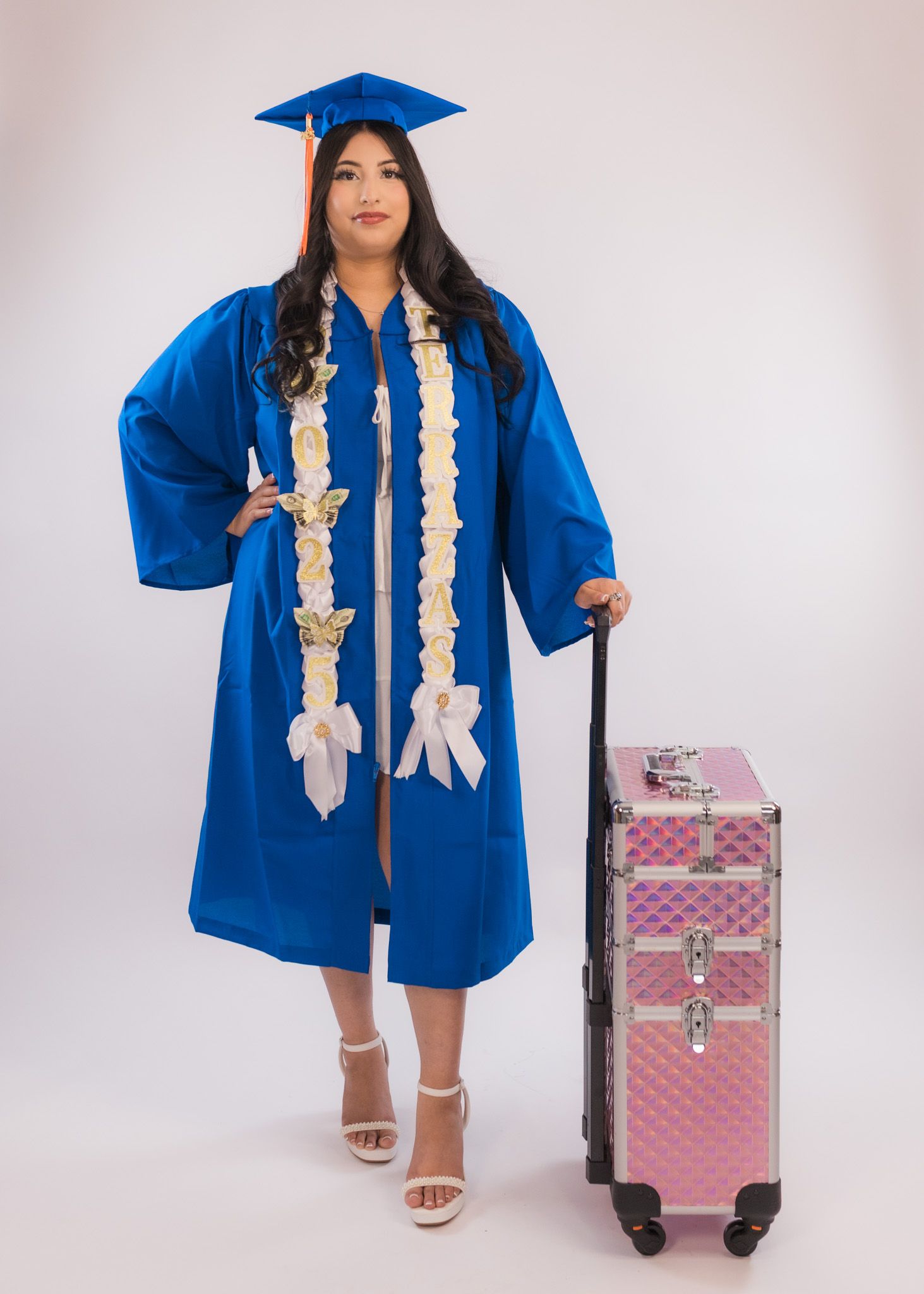 Graduate in blue cap and gown with white stole stands next to pink rolling suitcase.