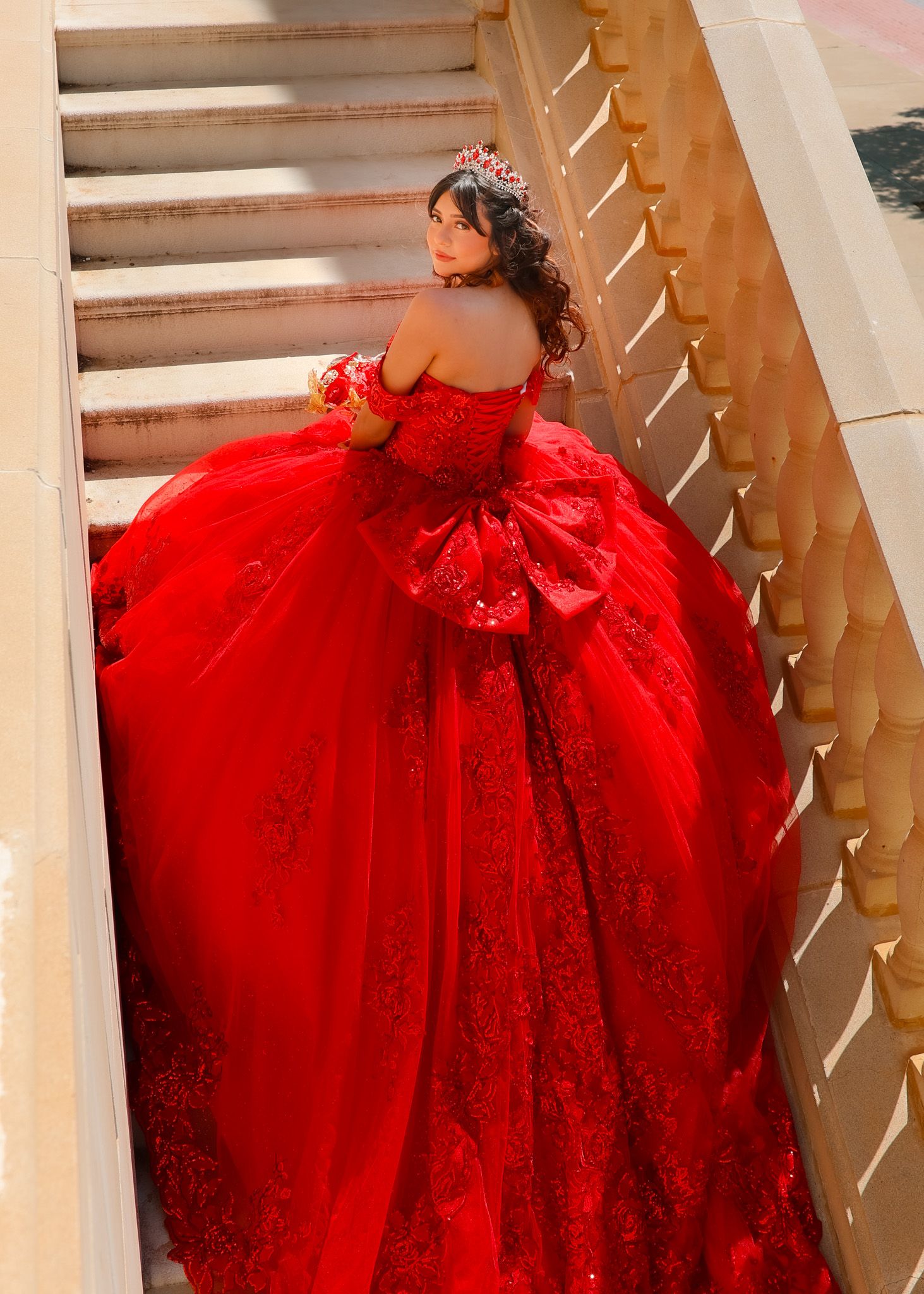 Woman wearing sparkly red quinceañera dress photographed from behind on elegant staircase.