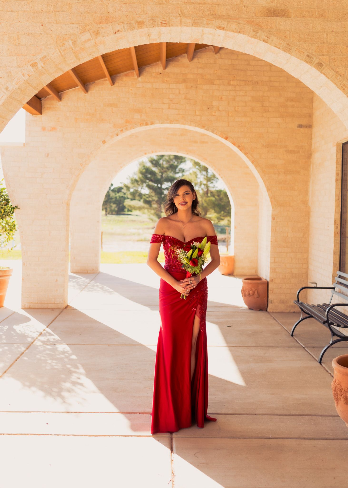Elegant formal portrait under arched walkway featuring red evening gown and bouquet.