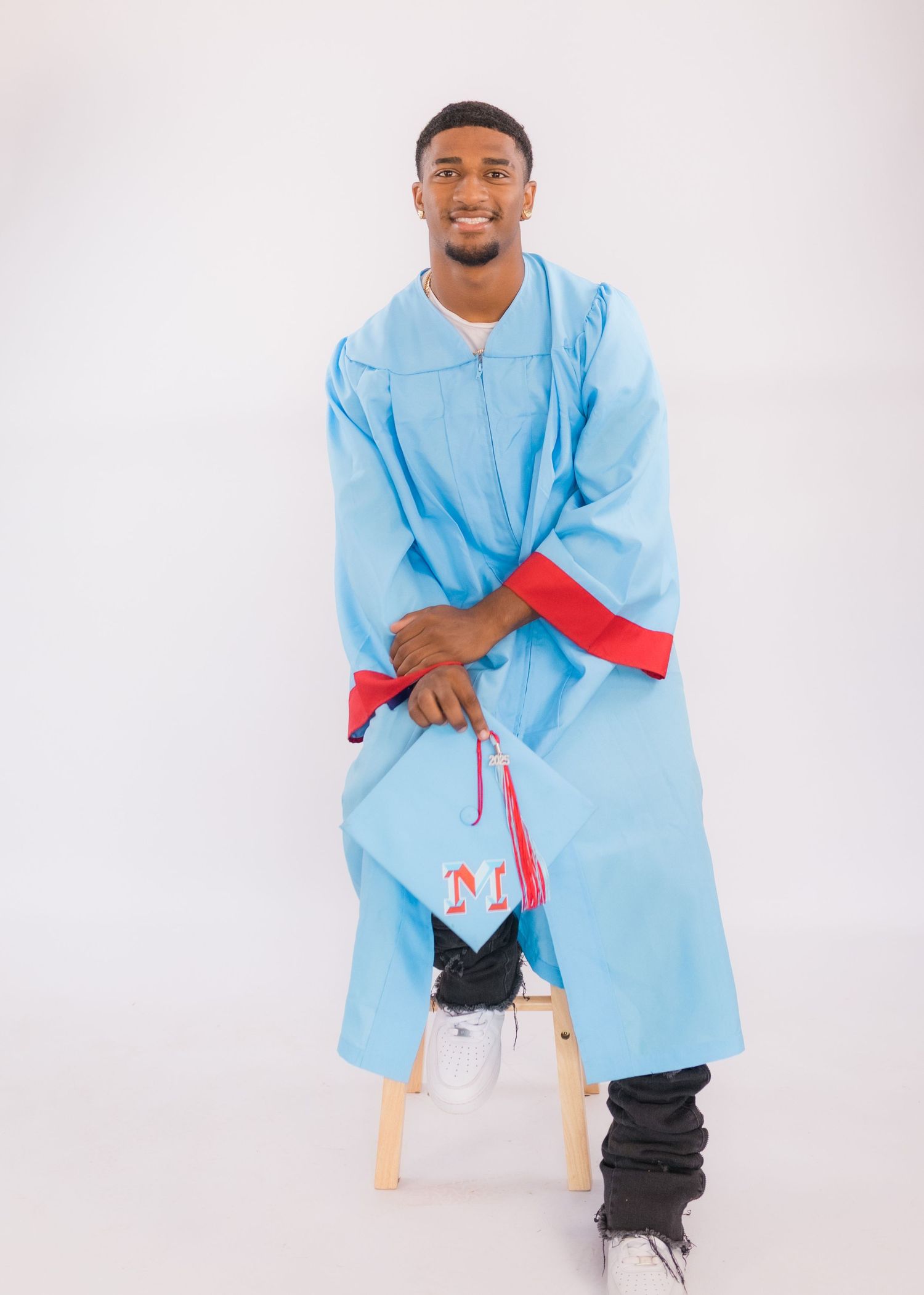 Graduate in light blue graduation gown sitting on stool against white background.
