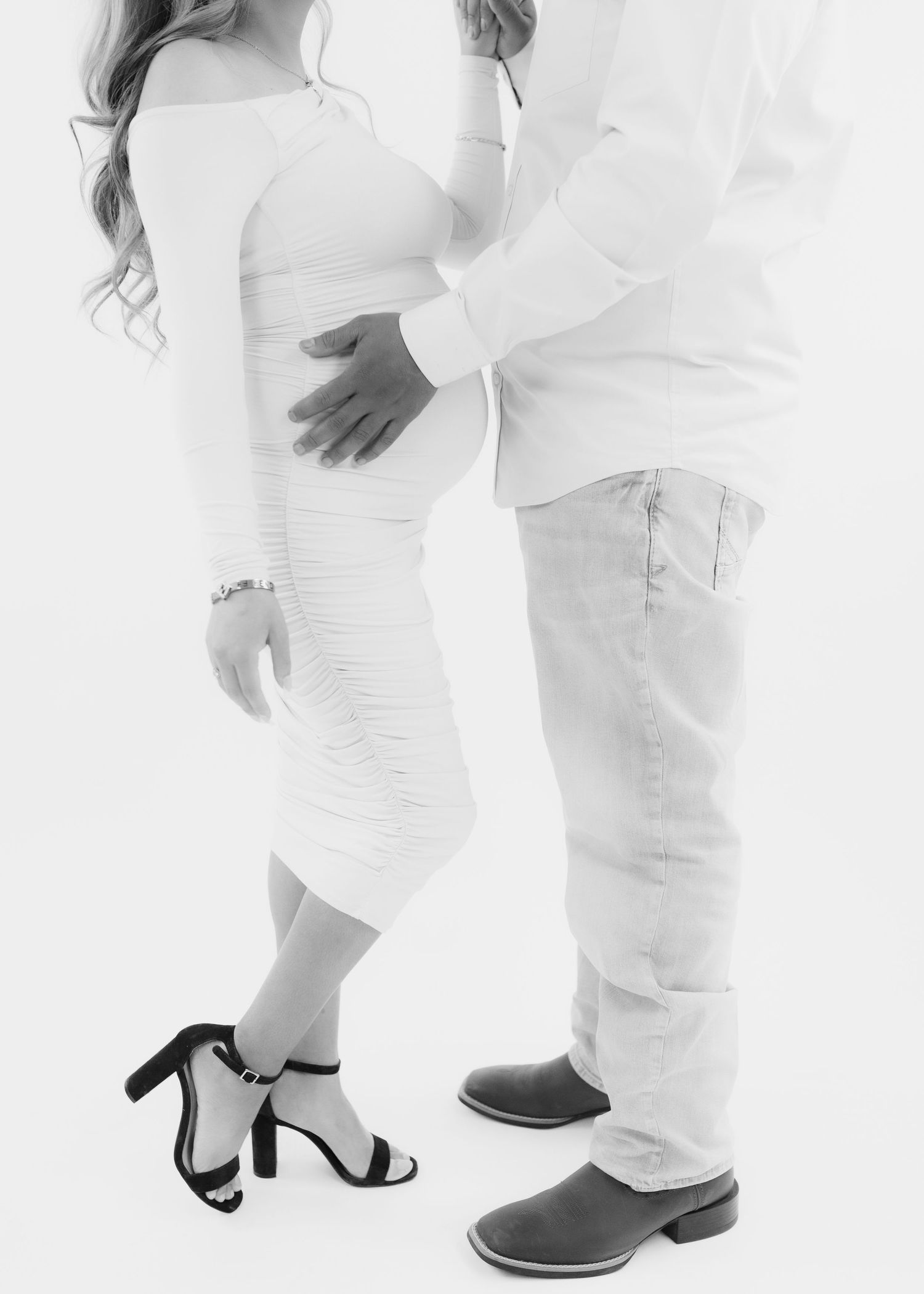 Black and white maternity photo of couple in white outfits, woman in heels, man in boots, embracing tenderly.