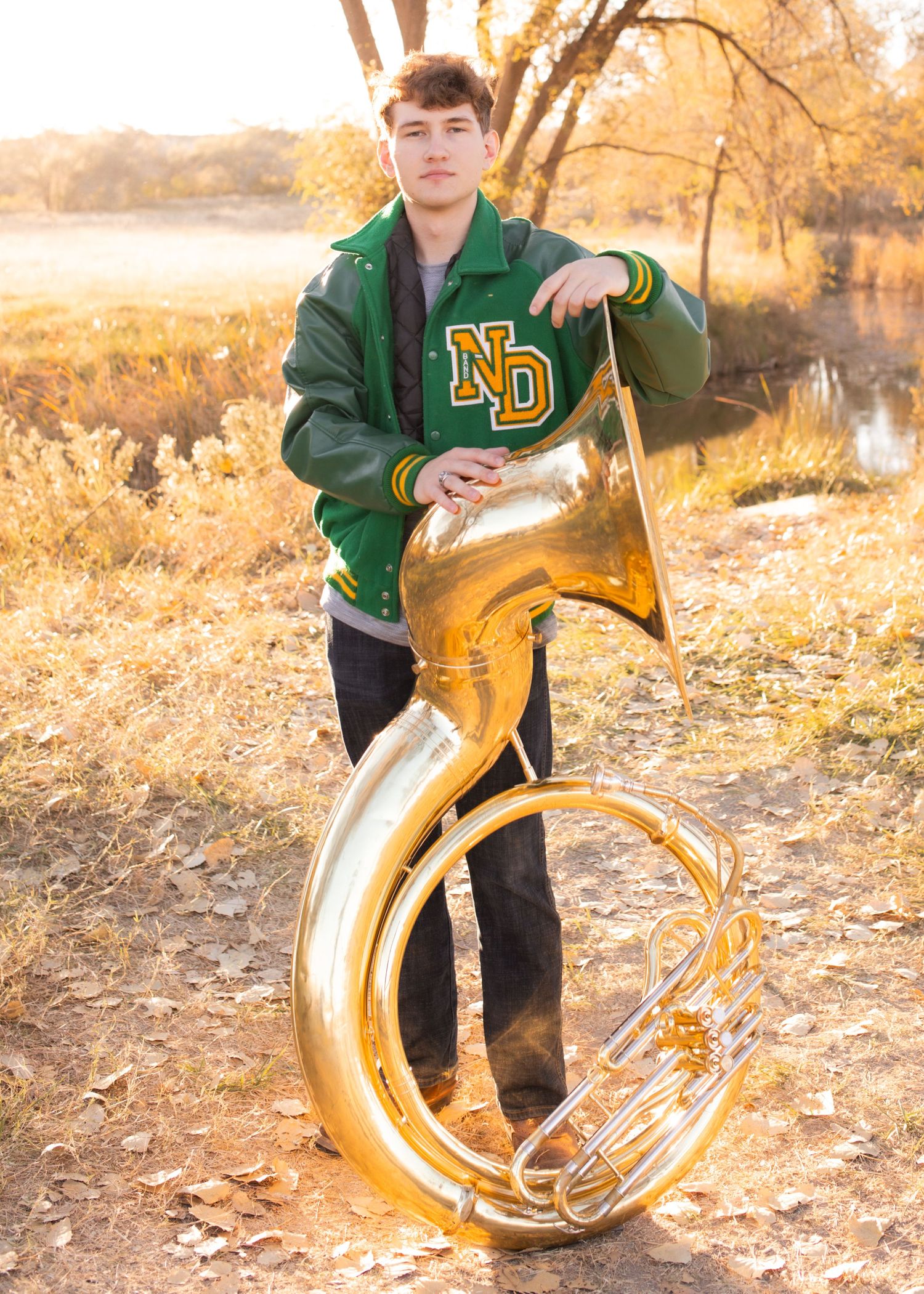 Young man in green ND varsity jacket poses outdoors with a shiny brass sousaphone in autumn setting.
