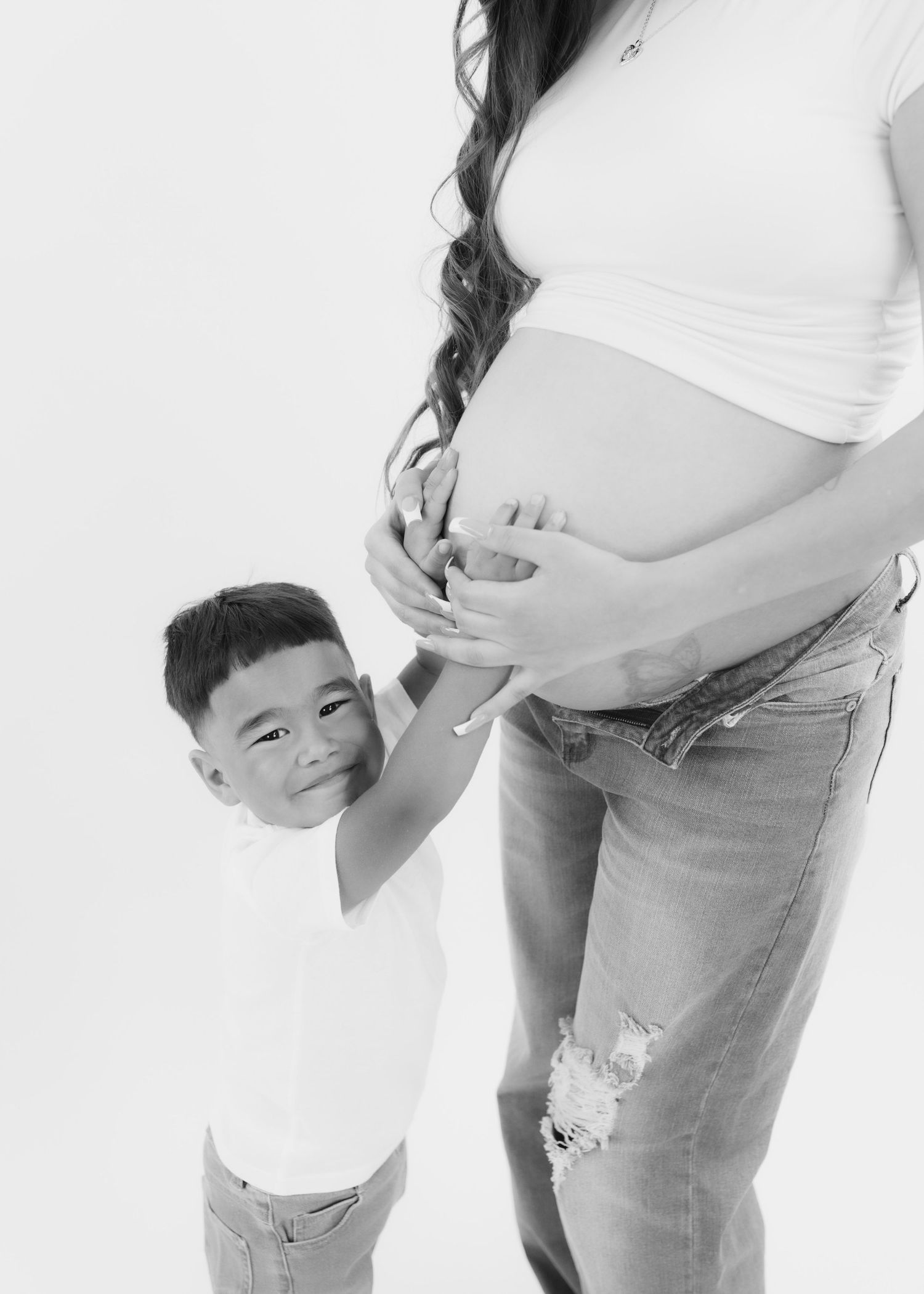 Black and white maternity photo of pregnant woman in jeans with young smiling boy touching her baby bump.