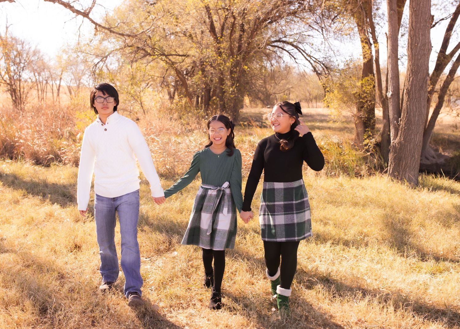 Three people holding hands walking through a golden autumn field with bare trees in the background.