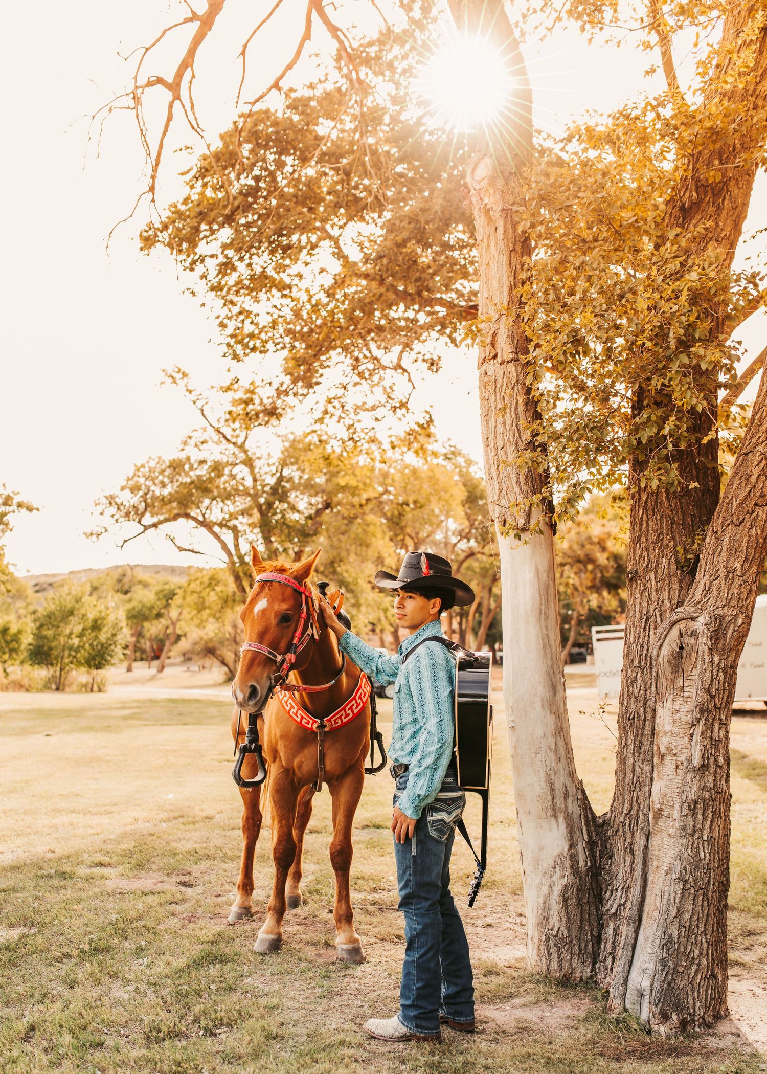 Cowboy in blue shirt standing with brown horse at sunset under trees.