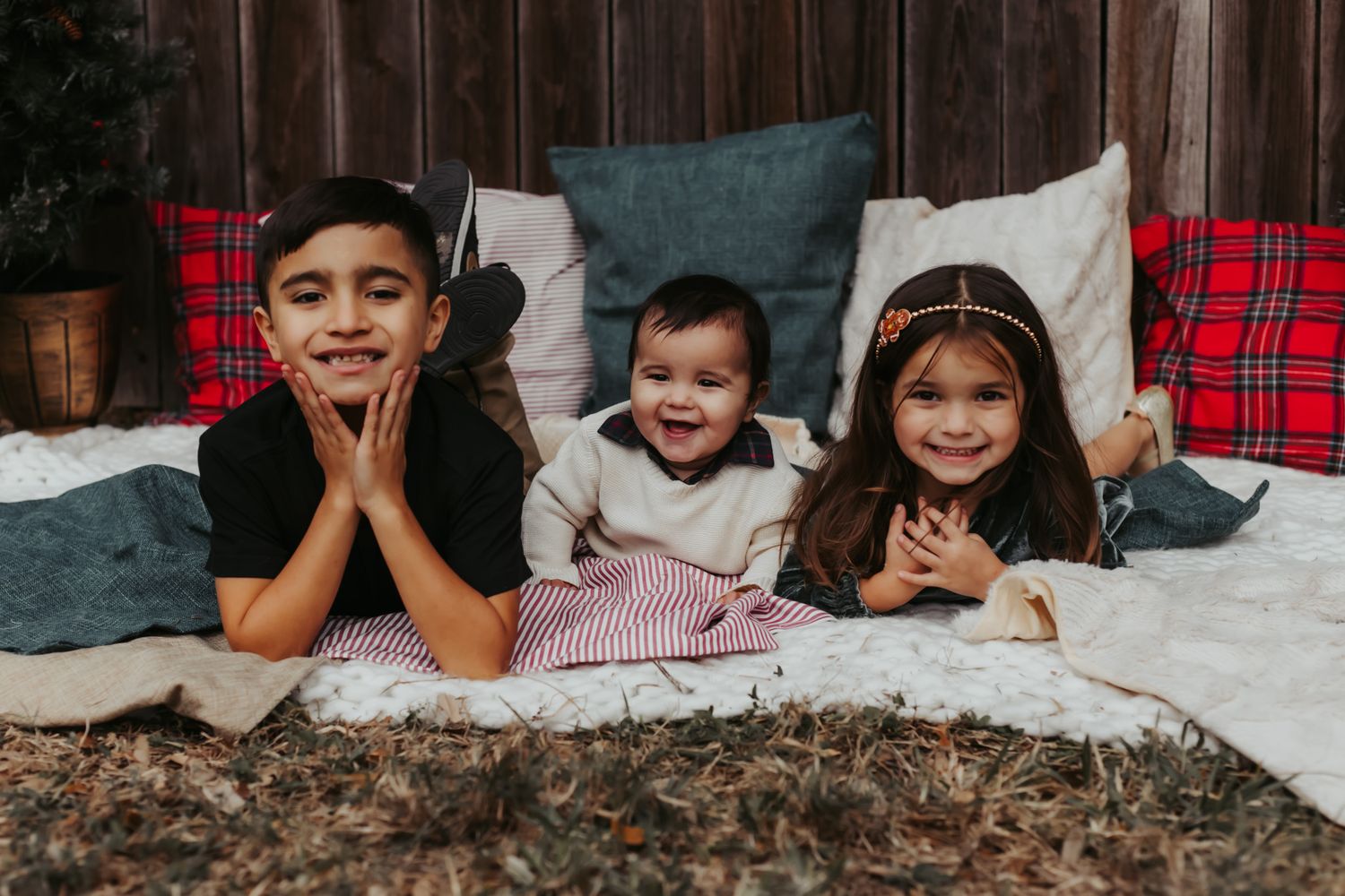 Three children lying on blankets with red plaid pillows against rustic wooden backdrop.