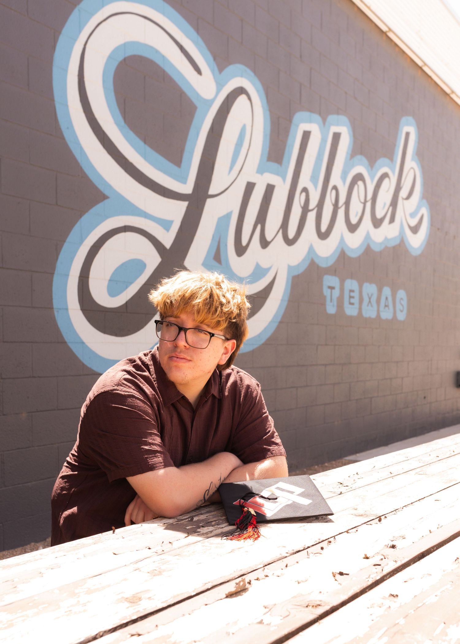 Young man with glasses sits at a wooden table in front of a Lubbock Texas mural on a brick wall.
