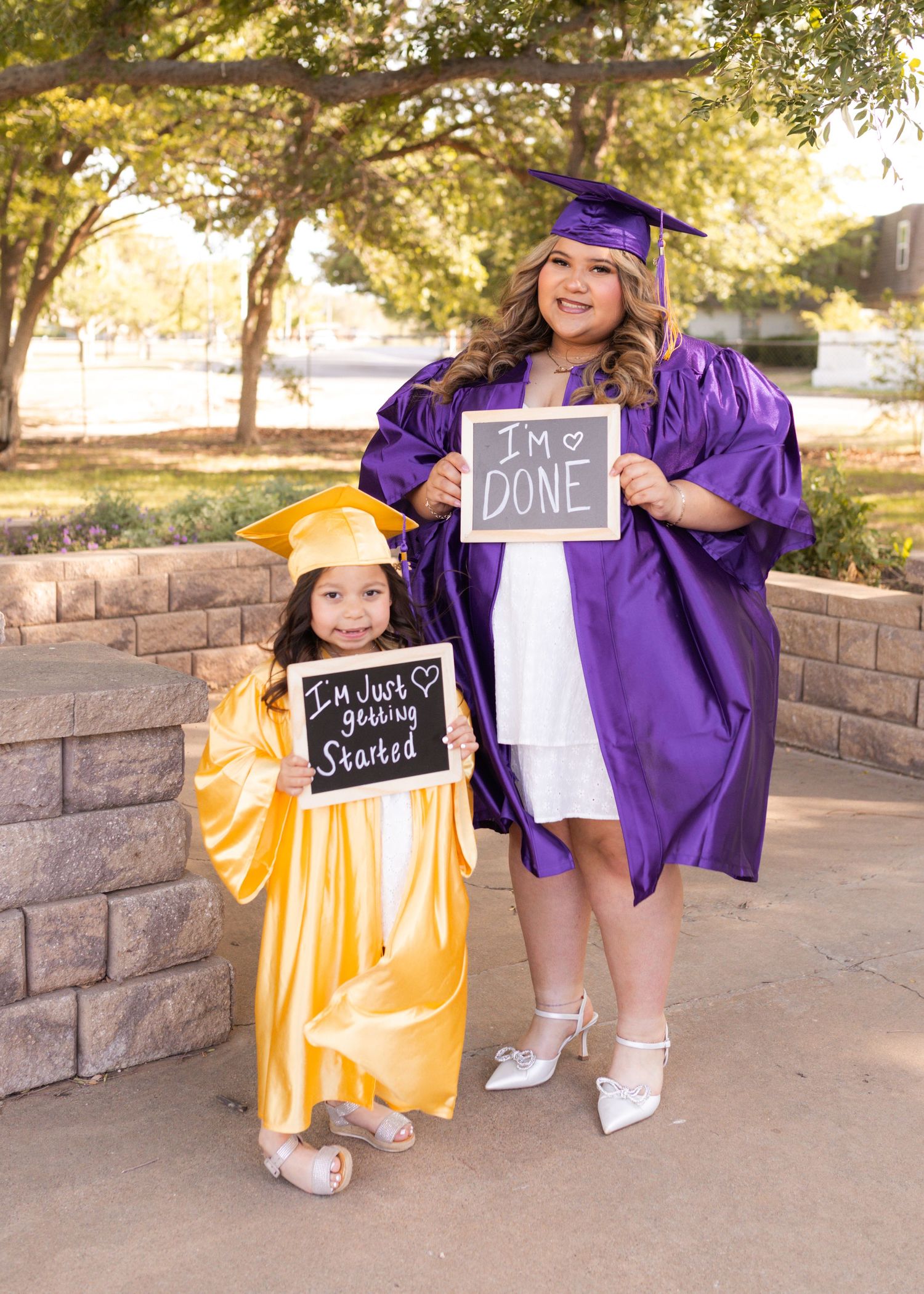 A young girl in gold cap and gown holds a sign saying I'm Just Getting Started beside a woman in purple graduation attire holding I'm Done.