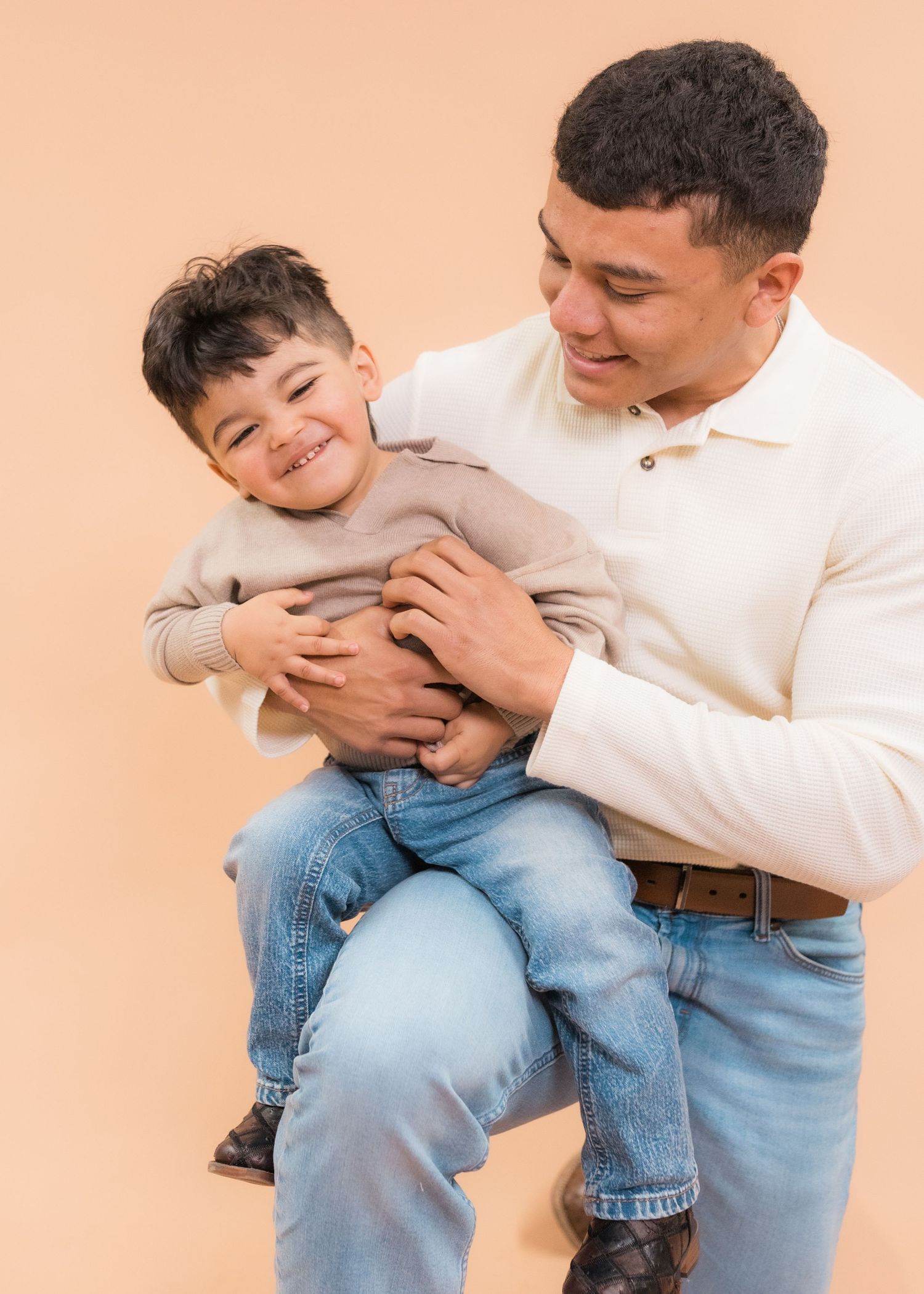 A smiling man holds a laughing young boy in his arms against a peach background.
