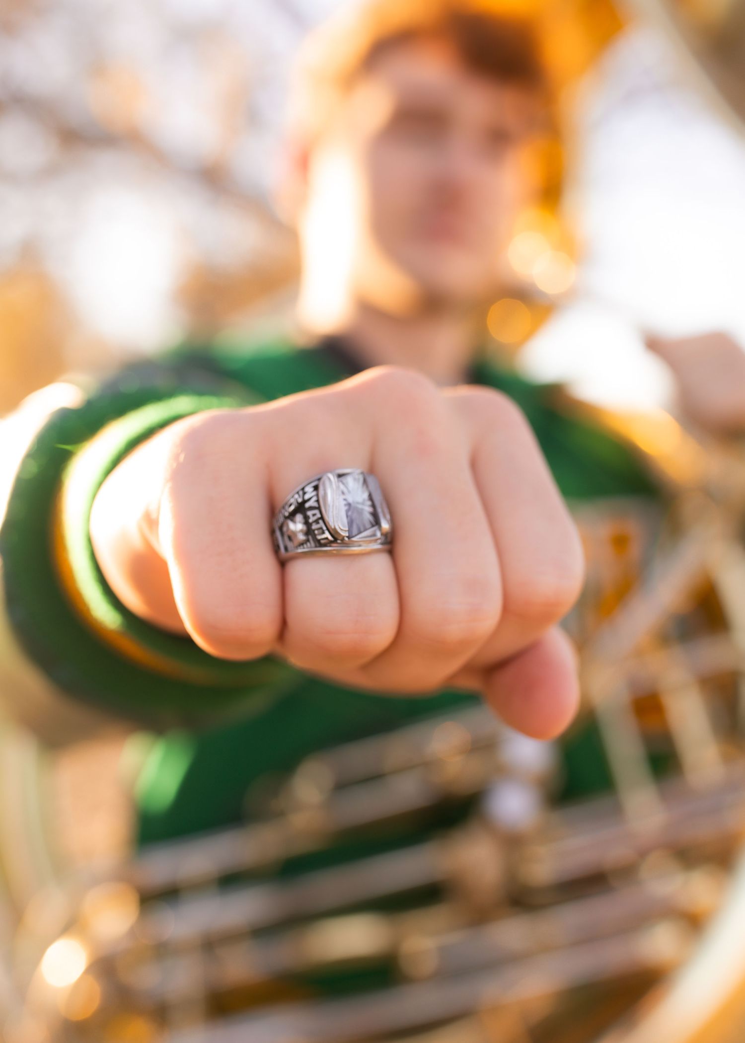A young man in a green sweater proudly displays a silver class ring with a large stone on his raised fist.